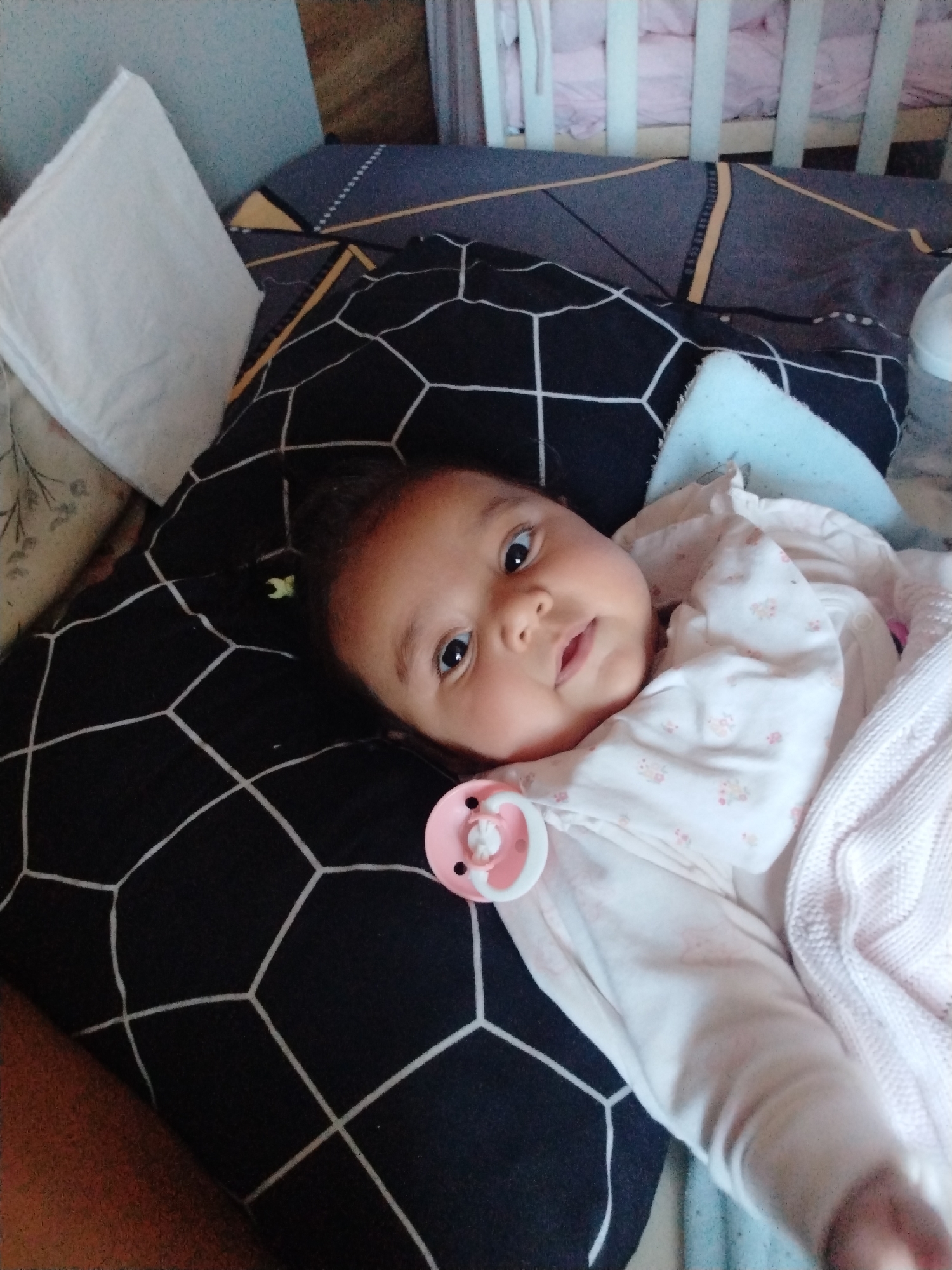 A baby is lying on a bed with a black and white patterned pillow. The baby has a pacifier in its mouth and is covered with a white and pink blanket. There is a white pillow on the left side of the image and a crib in the background. The location is unknown.