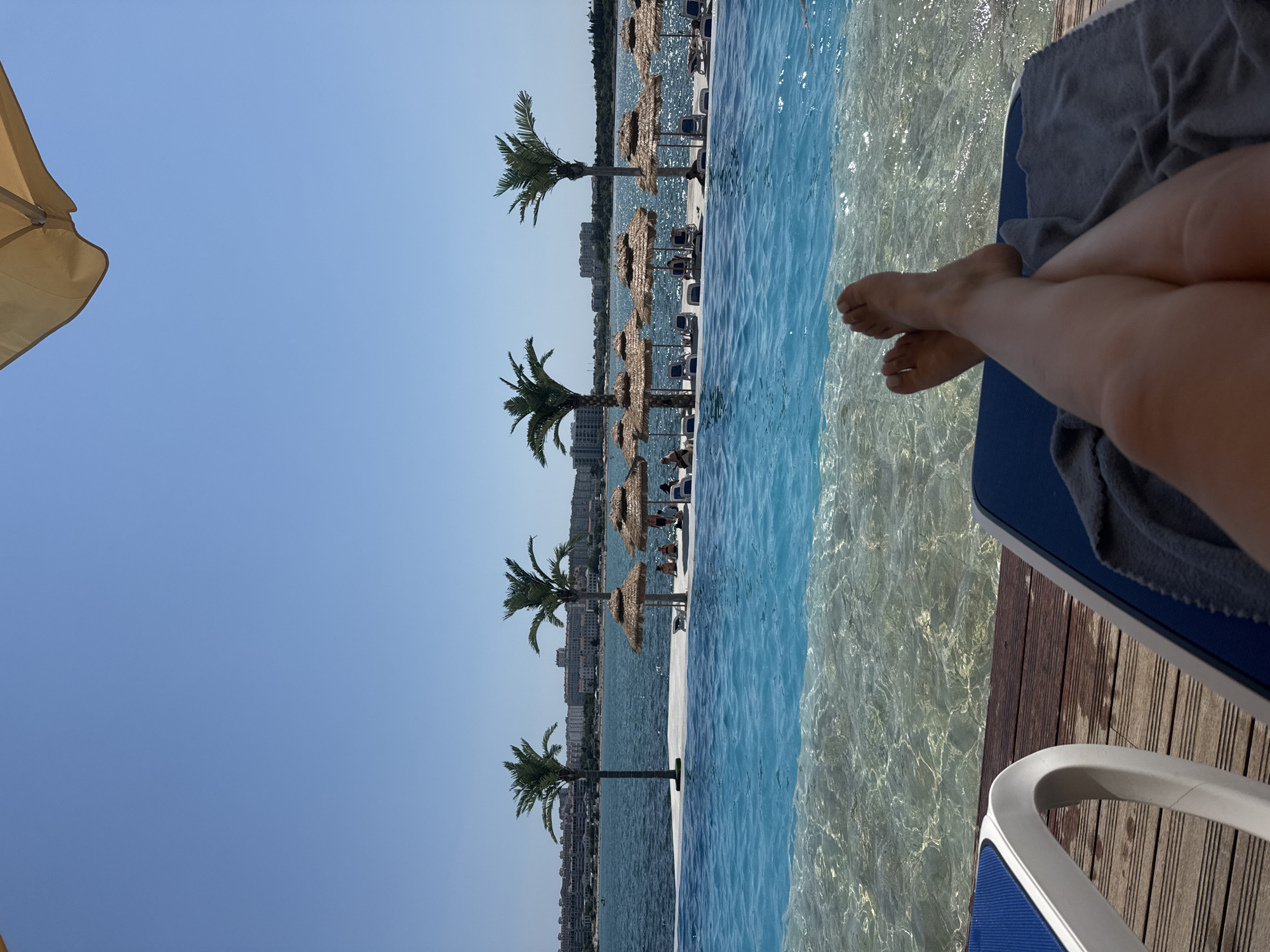 The image depicts a serene outdoor scene at a poolside resort. The foreground features a person's legs, crossed at the ankles, resting on a lounge chair. The chair is covered with a grey towel. Adjacent to this chair is another lounge chair with a blue seat and white frame. Both chairs are positioned on a wooden deck.
Beyond the deck lies an infinity pool, its crystal-clear turquoise water shimmering under the sunlight. The shallow end of the pool has a sandy or pebbled bottom, visible through the water. In the mid-ground, a white pool edge separates the infinity pool from a body of water, likely the sea, which is a deep blue.
Along the edge of the sea, there are several straw umbrellas providing shade for lounge chairs. Behind these, two artificial palm trees stand tall. Further in the distance, a city skyline with numerous buildings is visible, stretching along the coastline. The sky is a clear, bright blue with no clouds. The overall atmosphere is one of relaxation and vacation.