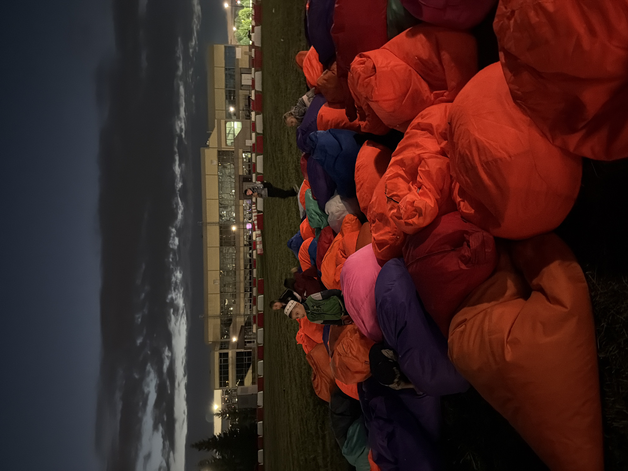 In an outdoor setting at dusk, a collection of large, brightly colored beanbag chairs are arranged on a grassy field. In the background, a modern, multi-story building with large windows is illuminated. Several children and adults are scattered among the beanbag chairs. One child, wearing a green jacket and a white and black patterned cap, stands prominently in the foreground, looking to the left. Another child is visible sitting in the beanbag chairs in the mid-ground, while an adult stands in the background near the building. The sky above is dark with some visible clouds. The scene appears to be a gathering or an event taking place in an open area, possibly a park or recreational space adjacent to a building.
