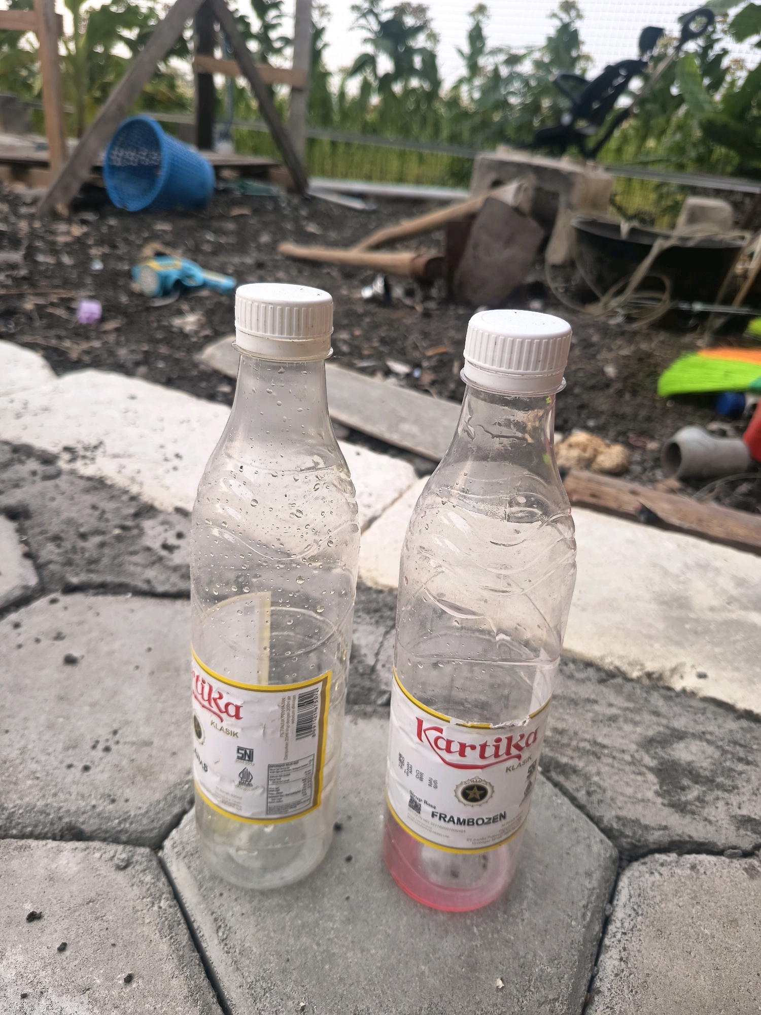 Two plastic bottles with white caps sit on a paved surface. The bottle on the left is mostly empty and has water droplets on its exterior. A label on the bottle reads "Kartika KLASIK". The bottle on the right contains a pink liquid and has a label that says "Kartika FRAMBOZEN KLASIK". The background is blurred and appears to be an outdoor area with some construction materials, including wooden beams and what looks like a blue basket. The scene is likely in Sidorejo, Indonesia, given the context.
