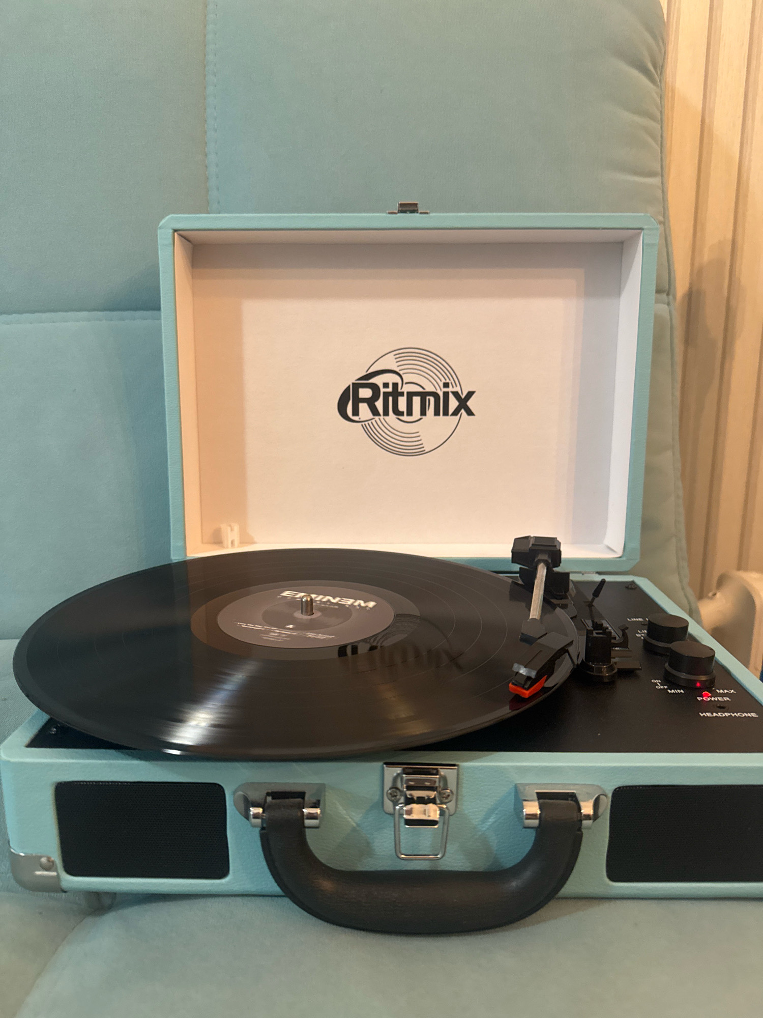 The image shows a light blue portable record player, open and with a black vinyl record playing on it. The record player is placed on a light blue upholstered surface, possibly a sofa or chair. The background consists of the same upholstered surface on the left and a vertically paneled wall on the right. The record player has a handle and metal clasps. The lid of the record player is open, revealing the brand logo "Ritmix" and an internal compartment. Controls for the record player, including knobs and a power indicator, are visible on the top surface. The scene is indoors. There are no people in the image. The location is not specified in the image or the provided context.