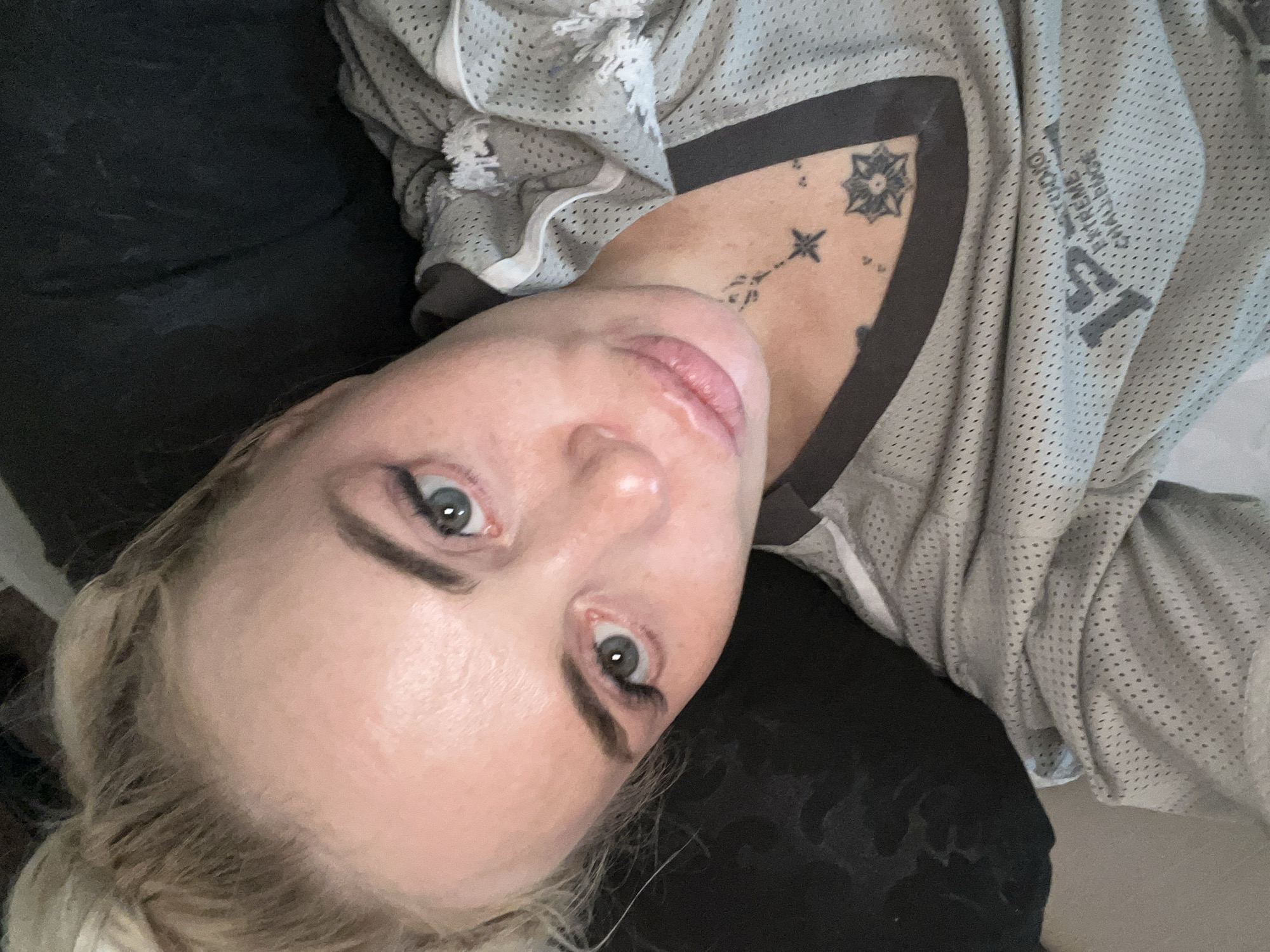 A selfie taken by a woman with blonde hair pulled up in a bun. She has green eyes, light skin, and light pink lipstick. She is wearing a grey mesh athletic jersey with white trim and black accents. A tattoo of a star and a celestial-like design is visible on her collarbone and upper chest. The background is dark and indistinct, suggesting an indoor setting. The overall impression is casual and personal.