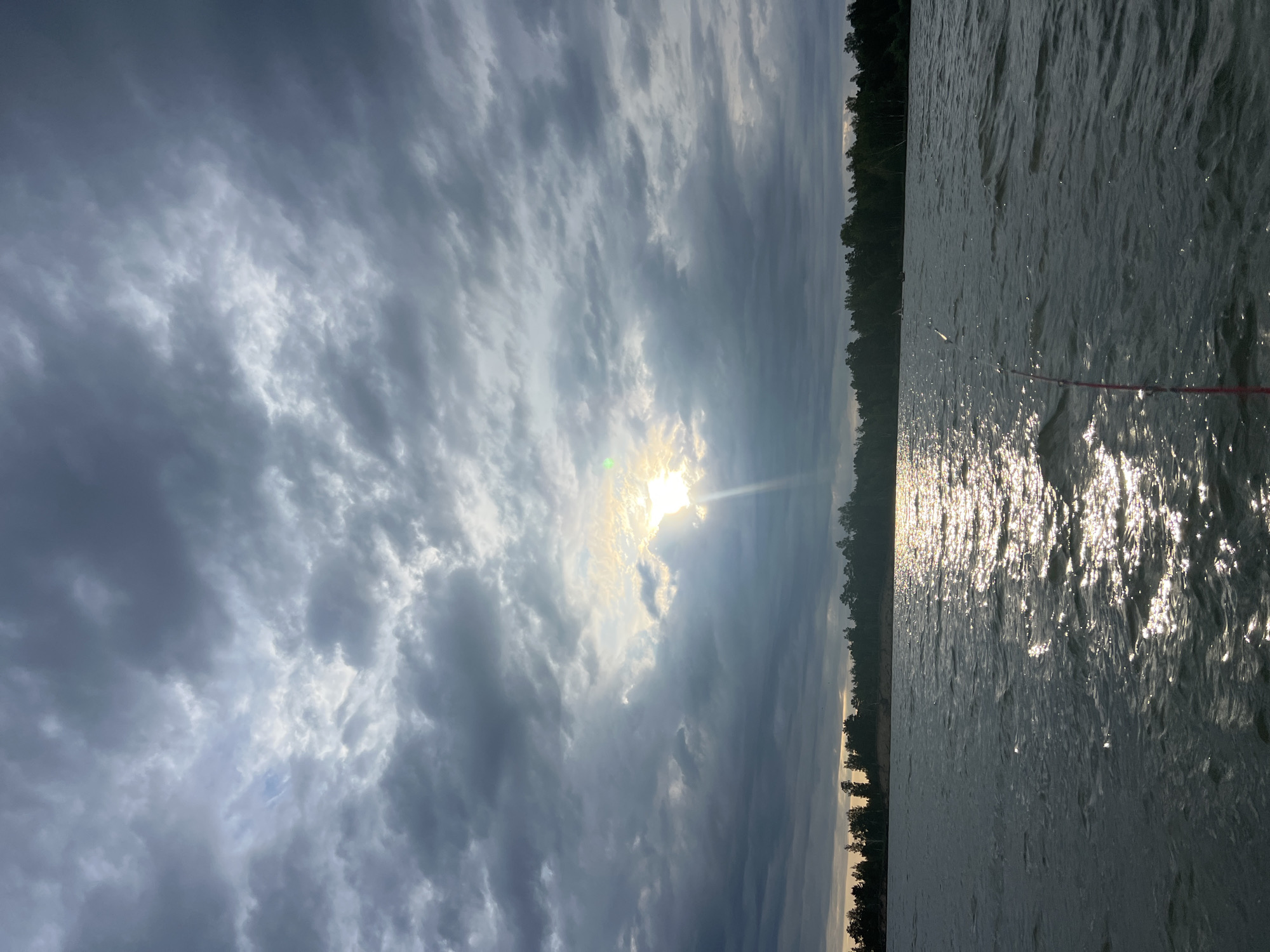 The image depicts a body of water, likely a lake, with a forest-lined shore in the distance. The sky is dramatic, filled with heavy, dark clouds partially illuminated by the sun breaking through. The sun's rays are visible, creating a bright glow and reflecting off the water's surface, which appears somewhat choppy. A fishing rod is visible in the lower right corner of the frame, extending into the water. There are no people or specific landmarks visible that would indicate the location as Borovoe, Russia.