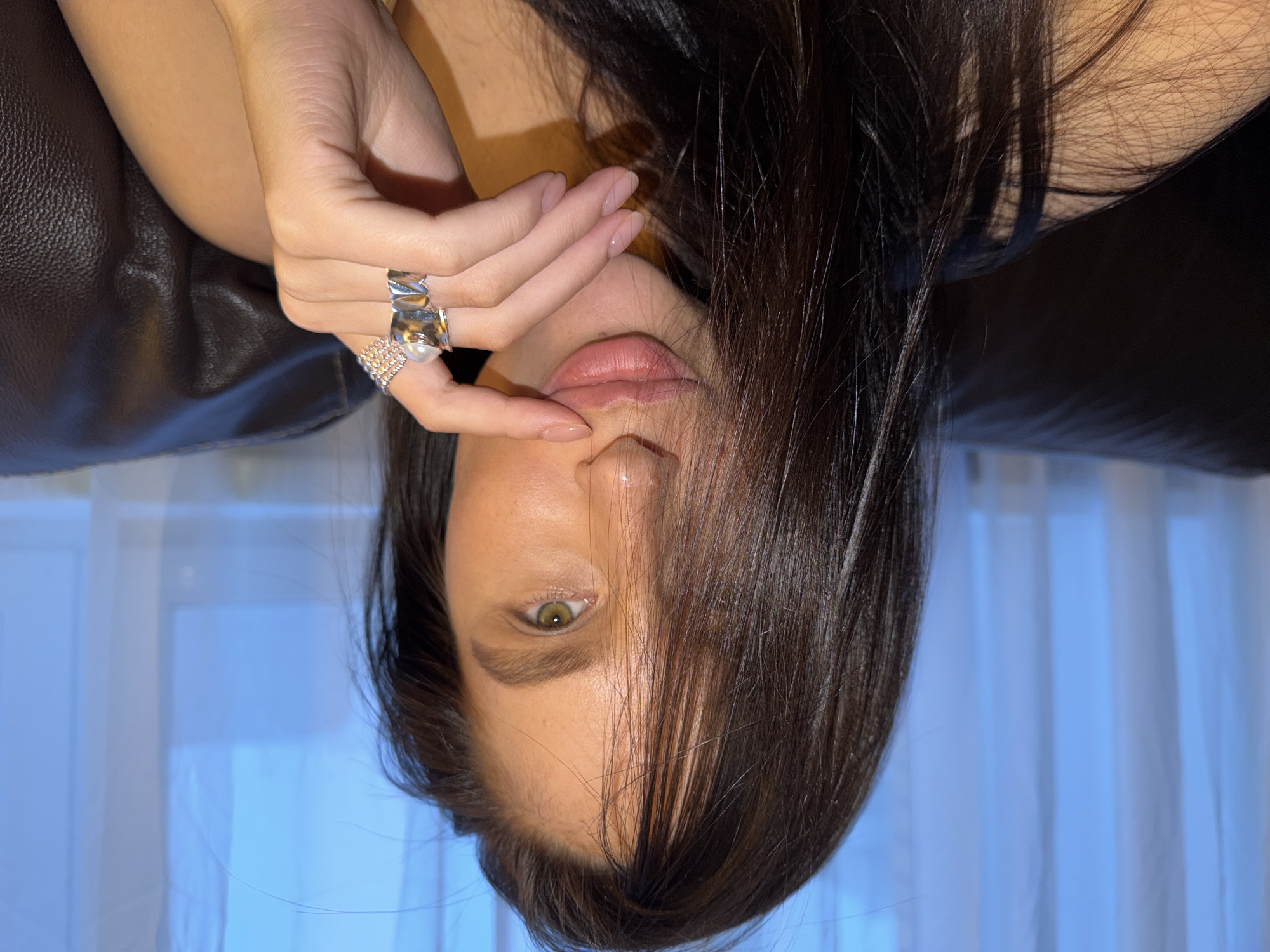 A close-up, portrait-style photograph shows a woman in a reclined or tilted position, with her dark brown hair flowing downwards and partially obscuring her face. Her head is angled, and her gaze, from a striking hazel or light green eye, is directed towards the viewer. Her dark eyebrows are well-defined.
Her right hand is raised, with fingers delicately touching her chin and lower lip. Her fingernails are neatly manicured with a natural, light pink polish. On her ring finger, she wears two prominent silver-toned rings: a wide, textured band featuring a large, spherical white pearl, and a narrower, pave-set ring with small, reflective elements below it. Her lips are a natural pink color and are slightly parted.
The background consists of a dark brown, textured surface on the upper left, possibly a leather sofa or chair, against which her arm rests. Behind her, a soft blue light emanates through translucent white curtains, indicating an indoor setting, possibly near a window. The lighting is soft and ambient, casting gentle shadows and highlighting her features. The overall impression is one of contemplation or relaxation. No text is visible in the image. The location context is Khimki, Russia.