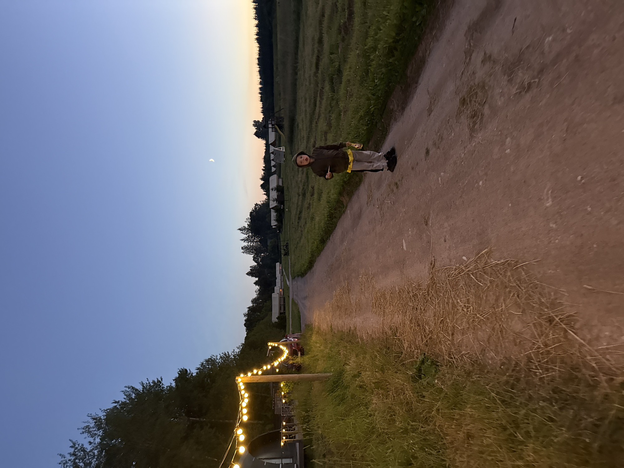 A young child stands on a dirt path at dusk, holding a sparkler. The child wears a dark hooded jacket and light-colored pants. The path is lined with tall grass and a string of illuminated bulbs on the left side of the image. In the background, there are several small buildings and a playground structure. The sky is a deep blue with a faint crescent moon visible. The scene appears to be in a rural or camping area.