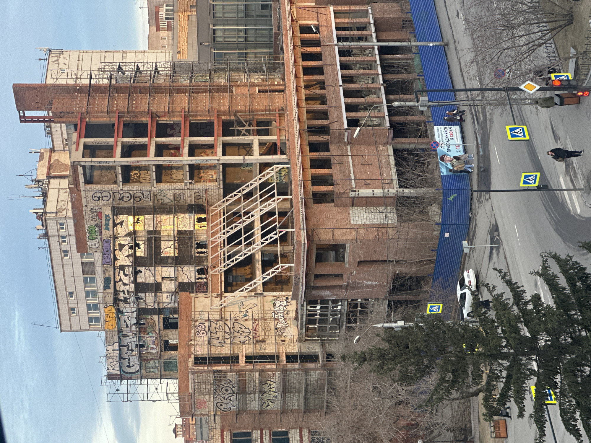 The image presents an urban street scene in Krasnoyarsk, Russia, centered on a large, multi-story building that appears to be either abandoned, under heavy renovation, or partially demolished. The structure features a mix of exposed brickwork and concrete, with many windows missing or boarded up. Extensive metal scaffolding covers much of the building's facade, which is heavily covered in graffiti, including numerous tags and Cyrillic script, such as "BBO" and letters that resemble "ОГРАБЛЕНО" (robbed).
The building borders a two-lane road, separated by a tall blue corrugated metal construction fence. Attached to this fence is a large billboard displaying a military recruitment advertisement. The billboard features images of people in military-style clothing and Russian text: "ПО КОНТРАКТУ" (by contract), "ВОЕННАЯ СЛУЖБА" (military service), and "ПРИСОЕДИНЯЙСЯ К СВОИМ" (join yours / join your own). A phone number "117" and website "служупоконтракту.рф" (servebycontract.rf) are also visible.
The street is equipped with standard urban infrastructure, including traffic lights, lampposts, and various Russian road signs such as pedestrian crossing signs, a 'yield' sign, and a 'stop' sign. A white car is parked along the roadside. Several pedestrians are seen walking on the sidewalk or crossing the street, including a lone person in a dark coat and a small group gathered near the traffic lights.
The time of day is daylight, with a clear or lightly clouded sky. The bare trees indicate late autumn or early winter weather. The overall impression is one of a functional city environment with a prominent derelict structure and a clear public messaging campaign.