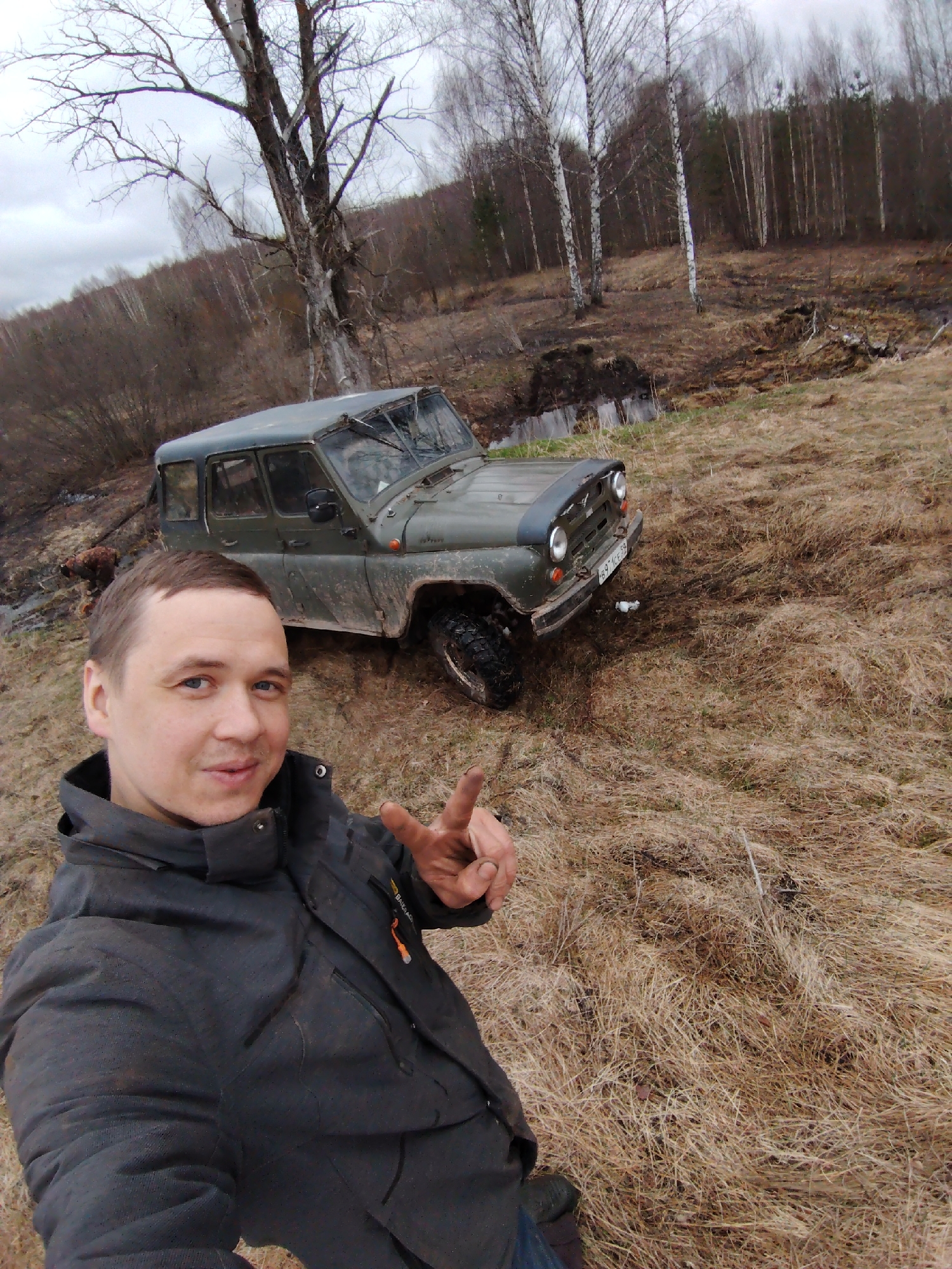 A man is taking a selfie in a rural setting in Markovskii, Russia. The foreground features a man wearing a dark jacket. He is making a peace sign. Behind him is a green, mud-covered UAZ-469 vehicle. The UAZ is partially stuck in mud, suggesting off-road activity. The background consists of a grassy field with dried vegetation. Bare trees and a dense forest line the horizon. The weather appears overcast. The location is near a small body of water or marshy area. The license plate is visible on the front of the vehicle, beginning with the number 69.