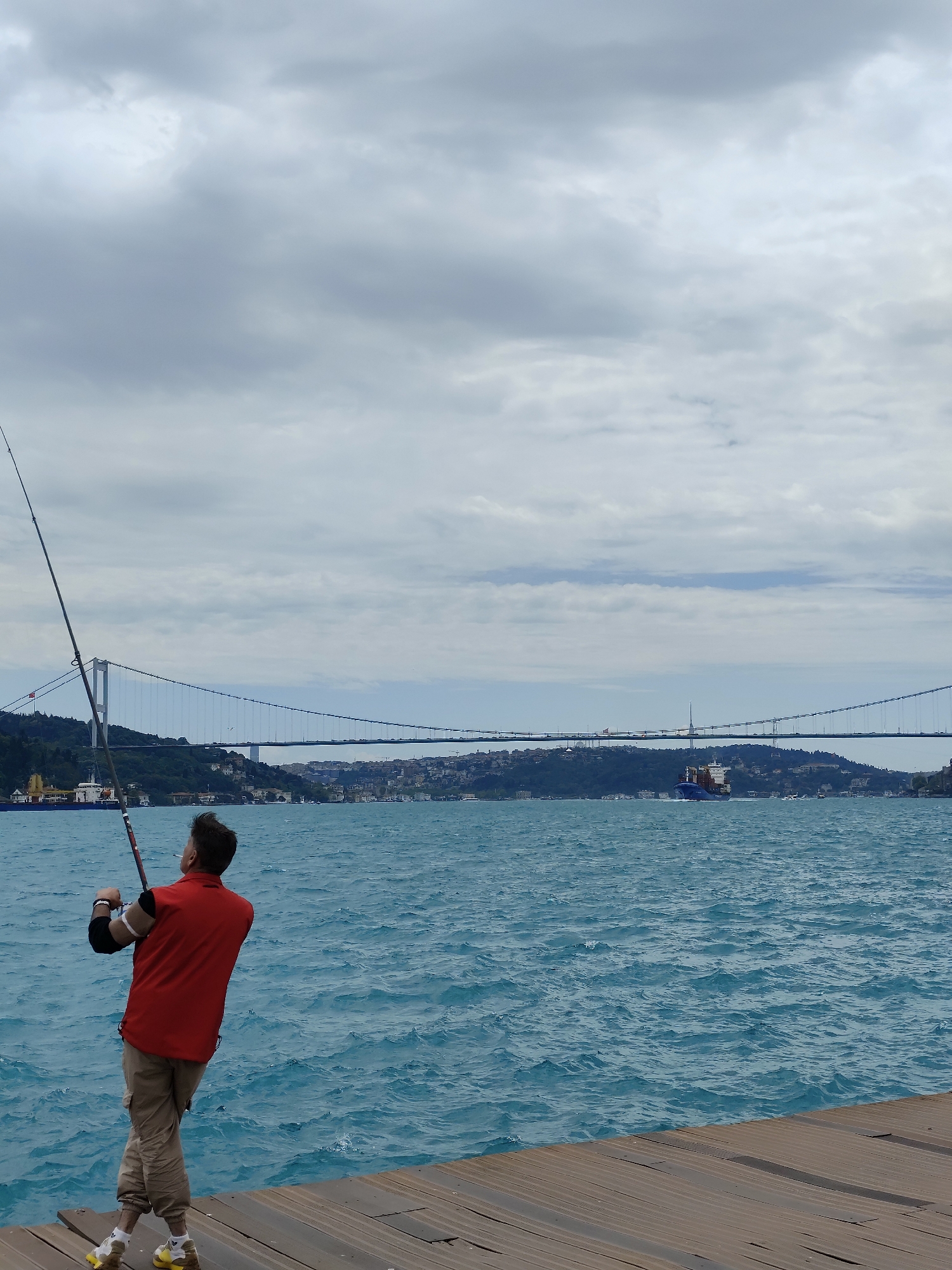 A man is fishing from a wooden dock. He is wearing a red shirt and khaki pants and holding a fishing rod, facing away from the viewer. In the background, the Bosphorus Bridge spans the water, with buildings and greenery visible on the opposite shore. A ship is sailing underneath the bridge. The sky is overcast with grey clouds. The location is in the city of Sarıyer, Turkey.