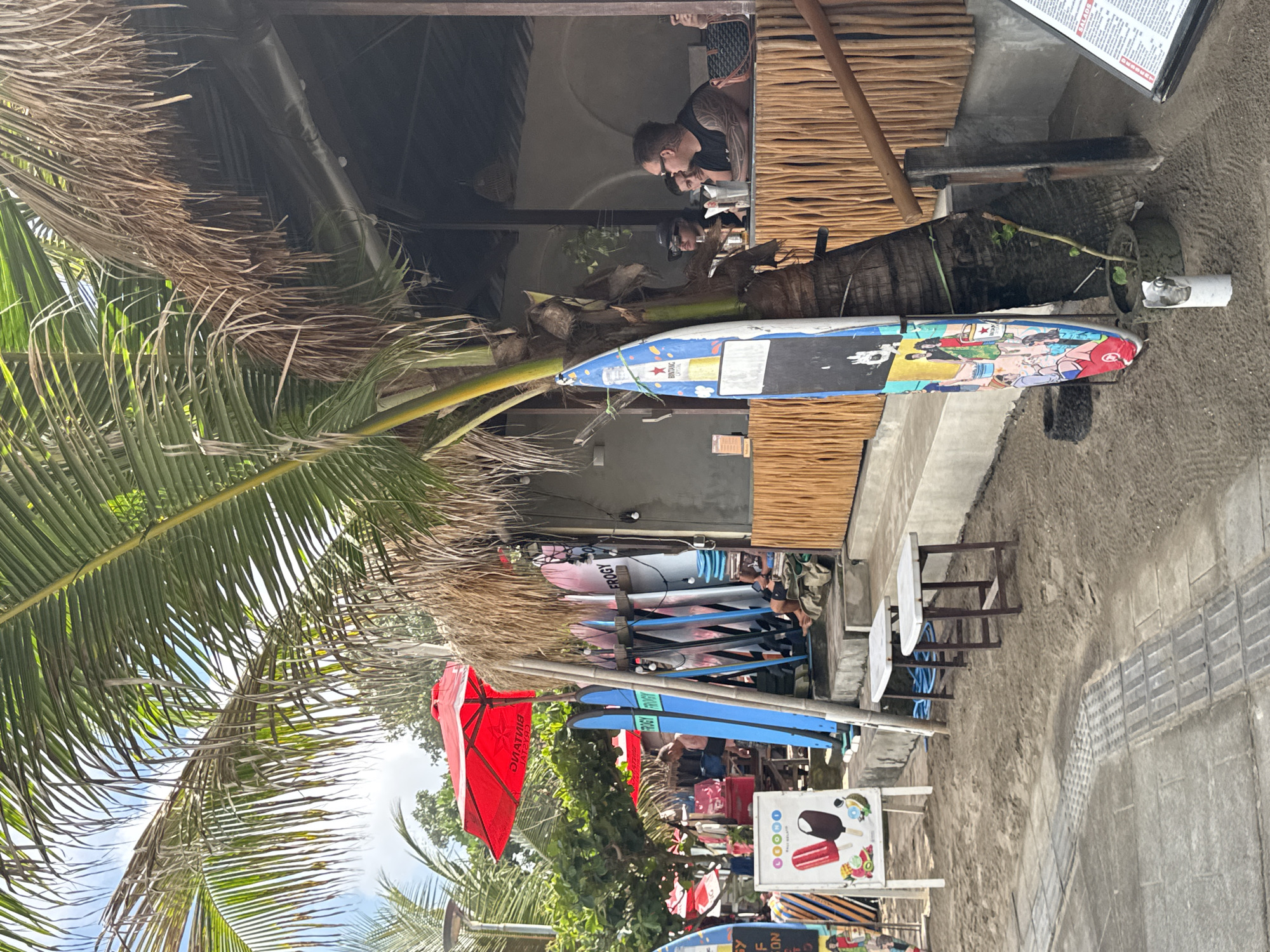 This image captures a vibrant, casual scene at what appears to be a beachside or roadside establishment, likely a cafe, restaurant, or surf shop, consistent with the Kuta, Indonesia location. The overall setting is sunny and tropical, indicated by the bright daylight and numerous prominent palm trees with both green and dried fronds.
Several people are visible. Inside a covered area with a thatched roof and wooden slat wall, a man with visible arm tattoos is seated at a table, seemingly dining or conversing with another person opposite him. Further along, more individuals are seen near racks of surfboards.
The scene is rich with objects. Numerous surfboards, varying in color and design, are propped up or stored on racks. One surfboard prominently features cartoon-like artwork and the brand "BINTANG," while others have "FROGY" visible. A large, bright red umbrella, also branded "BINTANG," provides shade outdoors. Various tables and chairs are scattered throughout, both inside the covered area and on the sandy/dirt ground outside. A white sign advertises "ICE CREAM" and "POPSICLE" with colorful illustrations of desserts and fruit. Another menu-like sign is partially visible, displaying "SALADS." The ground transition from concrete paving to a dirt or sandy area near the establishment.
The activity suggests a relaxed atmosphere, with people enjoying food or drinks and browsing surf-related items. The presence of surfboards, Bintang branding, and palm trees strongly reinforces the tropical, beach-oriented environment of Kuta.