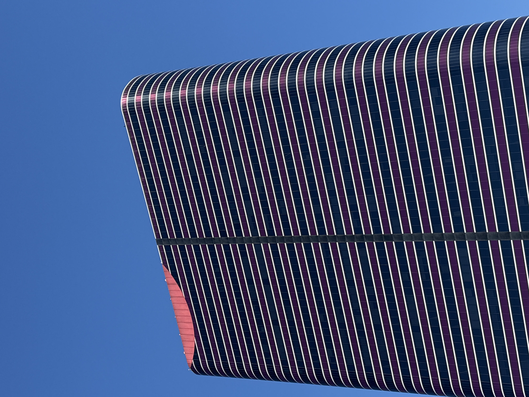 The image shows a close-up, low-angle shot of a modern, curved skyscraper with a distinctive facade against a clear blue sky. The building features alternating horizontal bands of dark purple and white. A vertical dark gray stripe runs down the center of the visible portion of the building. The top of the building has a red, curved accent. The image does not provide any information about the city of São Paulo, Brazil, or any people or actions.