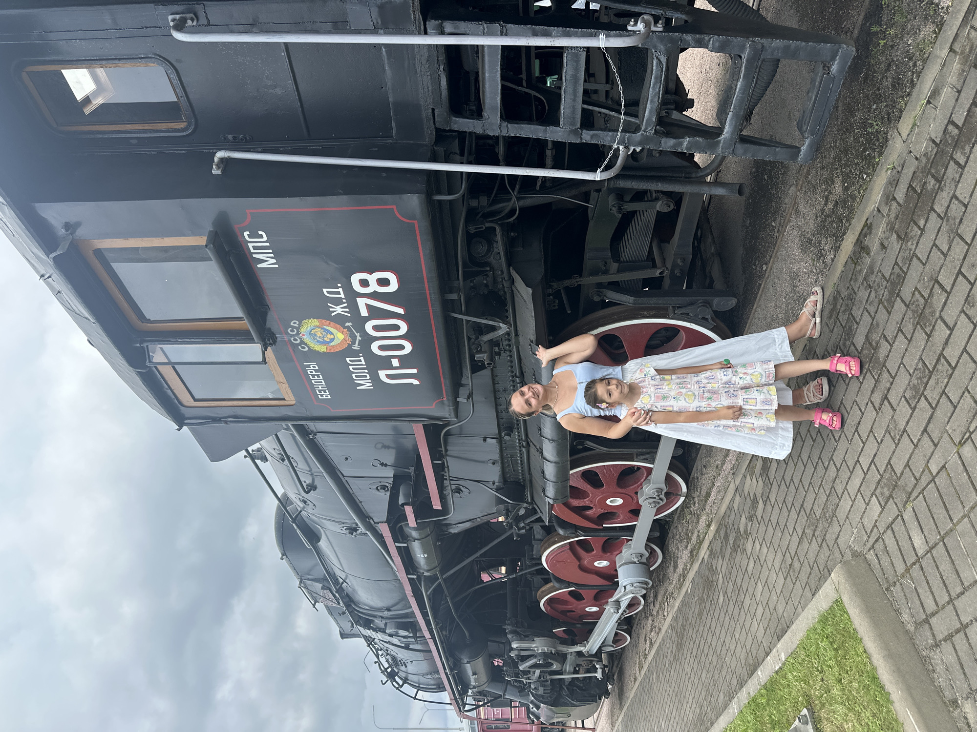 A woman and a young girl stand in front of a large, black steam locomotive. The woman is on the left, with her arm around the girl. She is wearing a light blue tank top and a long white skirt. The girl is wearing a colorful dress with a pattern of various cartoon characters. Both are smiling at the camera. The locomotive is a prominent subject, with large red wheels and visible mechanical details. Text is visible on the side of the locomotive, including Cyrillic letters and the number "Л-0078". The scene appears to be outdoors, with a paved walkway in the foreground and a cloudy sky above. The location is identified as Saint Petersburg, Russia, specifically at the Central Naval Museum.