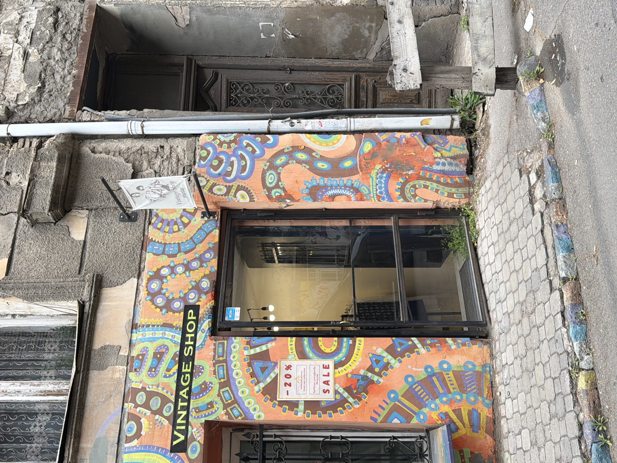 The image is of the exterior of a vintage shop in Tbilisi, Georgia. The shop's facade is painted in a vibrant, colorful, psychedelic pattern of orange, blue, and green. Above the shop window is a sign that reads "VINTAGE SHOP" vertically in black letters on a yellow background. The shop window displays a "SALE -20%" sign. The shop is located in an old building with weathered stone and exposed brick. A white pipe runs horizontally above the shop window. The sidewalk is paved with grey bricks.