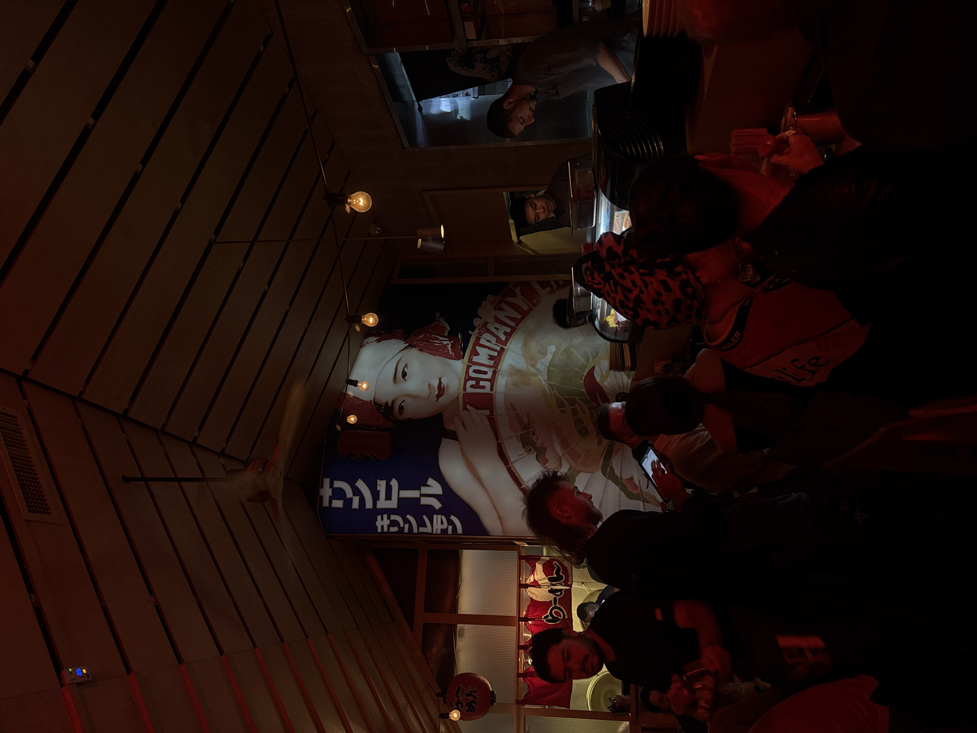 The image shows the interior of a bar or restaurant in Tel Aviv, Israel. The space features a striking vintage-style advertisement on the wall, depicting a woman holding a product labeled "Company" in English and Hebrew. The restaurant is populated with customers, some seated at the bar, others standing. The ambiance is dim, with exposed light bulbs providing a warm glow. The decor includes wooden slats on the walls and ceiling, and traditional Japanese lanterns are visible. The scene suggests a lively, social atmosphere.