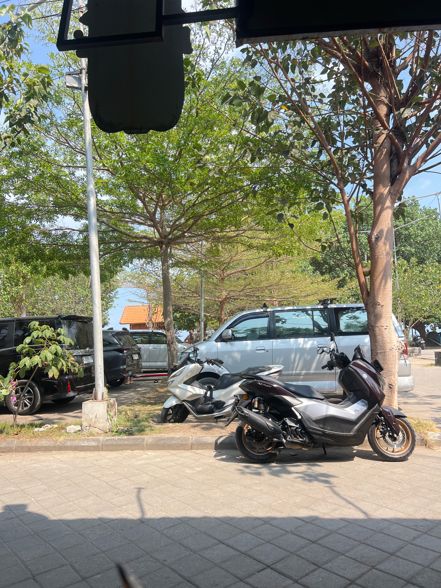 In Kuta, Indonesia, a sunny outdoor scene depicts a paved area with several parked vehicles. In the foreground, two scooters are parked, one white and silver, the other dark maroon and silver. Behind them, a silver minivan is parked, and to its left, a black SUV is partially visible. A tall utility pole stands between the SUV and the minivan. Lush green trees with visible branches and foliage are scattered throughout the scene, casting shadows on the ground. A building with an orange tiled roof is visible in the background, and a distant view of the ocean under a clear blue sky can be seen beyond it. The overall environment suggests a warm, tropical setting, likely a parking area near a beach or coastal establishment.
