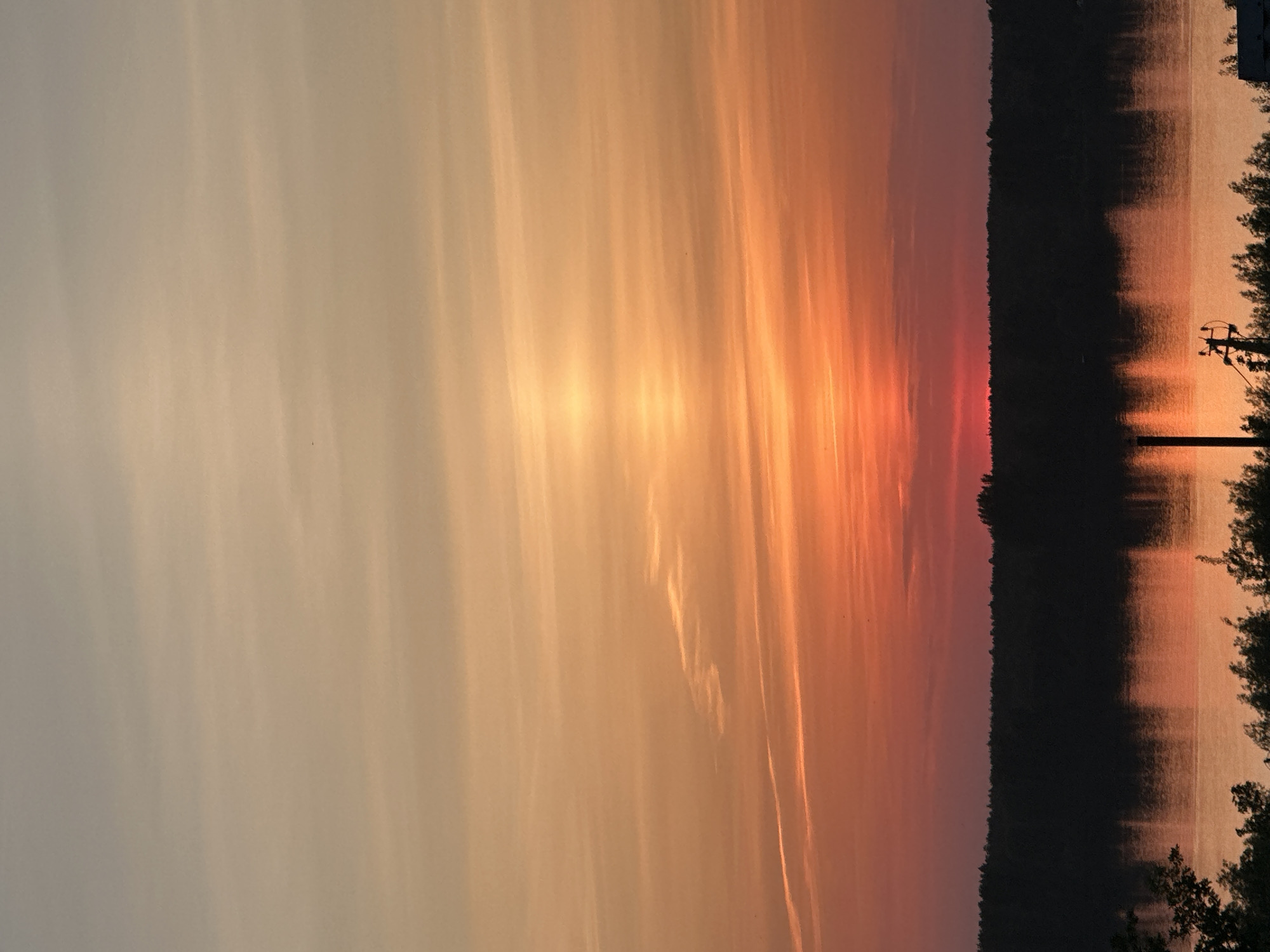 The image depicts a sunset over a body of water, possibly a lake or river, next to a treeline. The sky is filled with orange and pink hues from the setting sun. The silhouette of trees and a utility pole is visible on the right side of the image, reflected in the water. The location is unknown as information regarding Chaikovsky, Russia is not explicitly stated in the image.