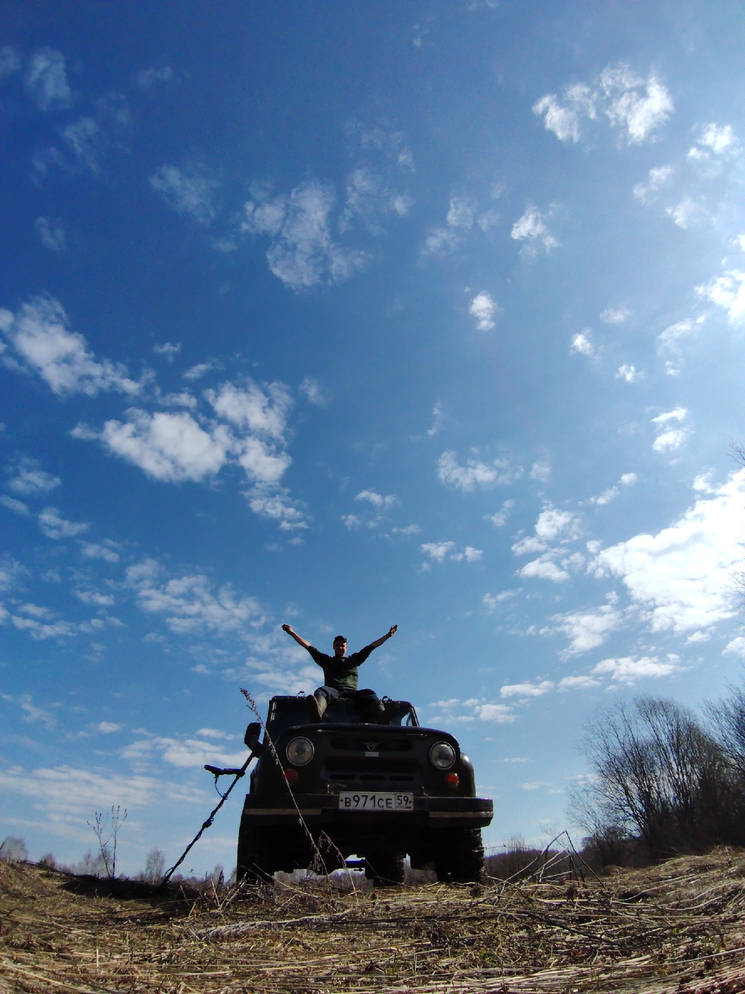 A high-angle, wide shot captures a person sitting triumphantly on the hood of a dark, rugged off-road vehicle under a vast, bright blue sky in Markovskii, Russia. The individual, a male, wears a dark top and is partially silhouetted against the bright sky, with his arms outstretched in a gesture of freedom or joy. His face is visible, showing a slight smile.
The vehicle is parked on dry, uneven ground covered with sparse, light-brown vegetation and scattered dead branches, indicative of late winter or early spring. A long, dark object, possibly a metal detector or a pole, leans against the left front side of the vehicle. The front of the vehicle, including its grille, circular headlights, and part of the bumper, is visible. A Russian license plate, "B971CE 59", is clearly affixed to the front bumper.
The sky dominates the upper two-thirds of the frame, featuring scattered white cumulus clouds. A bright sun is visible in the upper right corner, casting a strong light and creating a slight lens flare. In the far background on the right, there are some bare trees and bushes, suggesting a rural or wilderness setting. The overall scene conveys a sense of outdoor adventure and exhilaration on a sunny day.