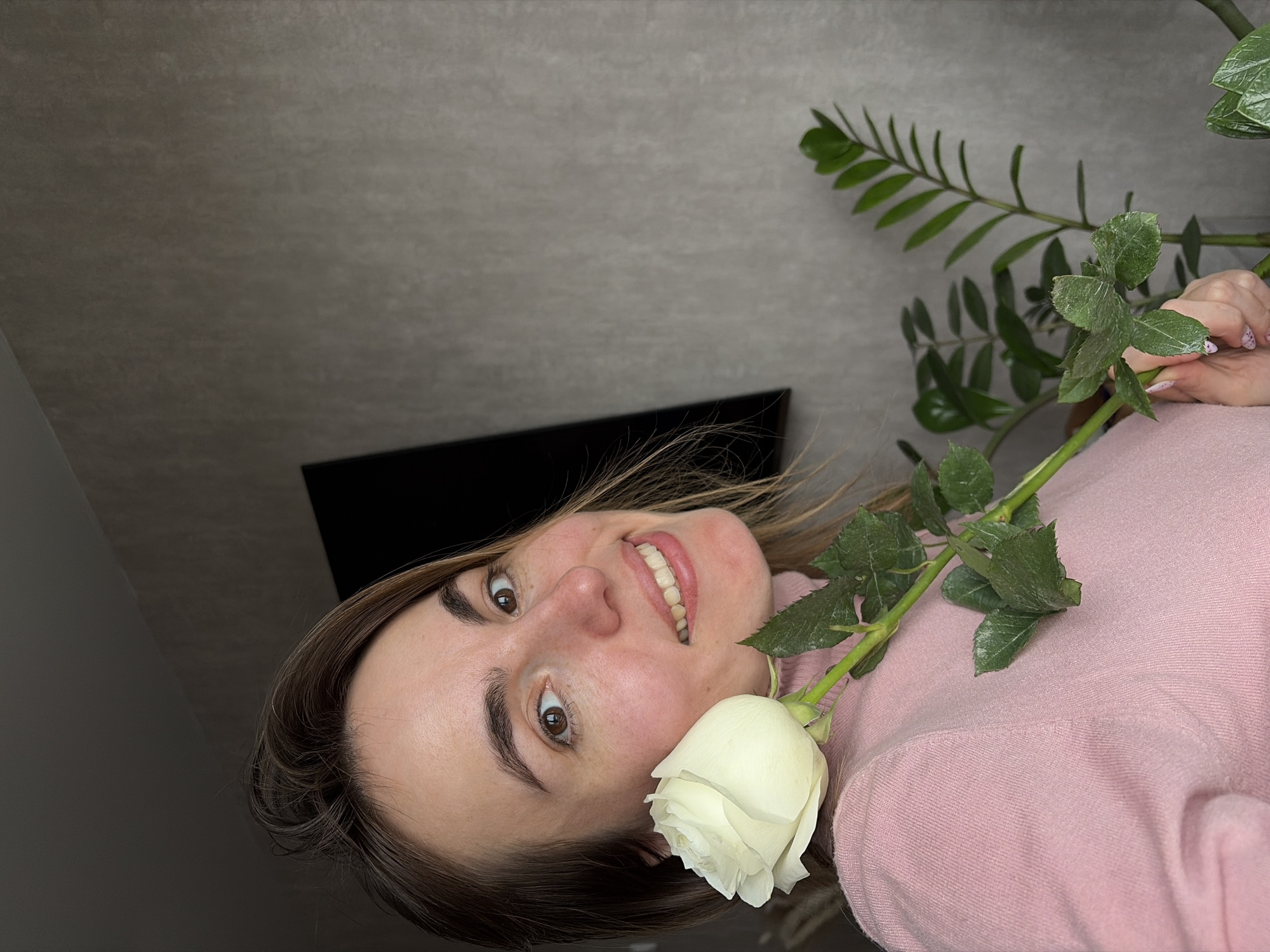 A woman is holding a white rose in front of her, smiling at the camera. She is wearing a pink sweater. Behind her is a green leafy plant and a gray wall. A black object is also visible in the background. There is no indication of the location being Izhevsk, Russia.