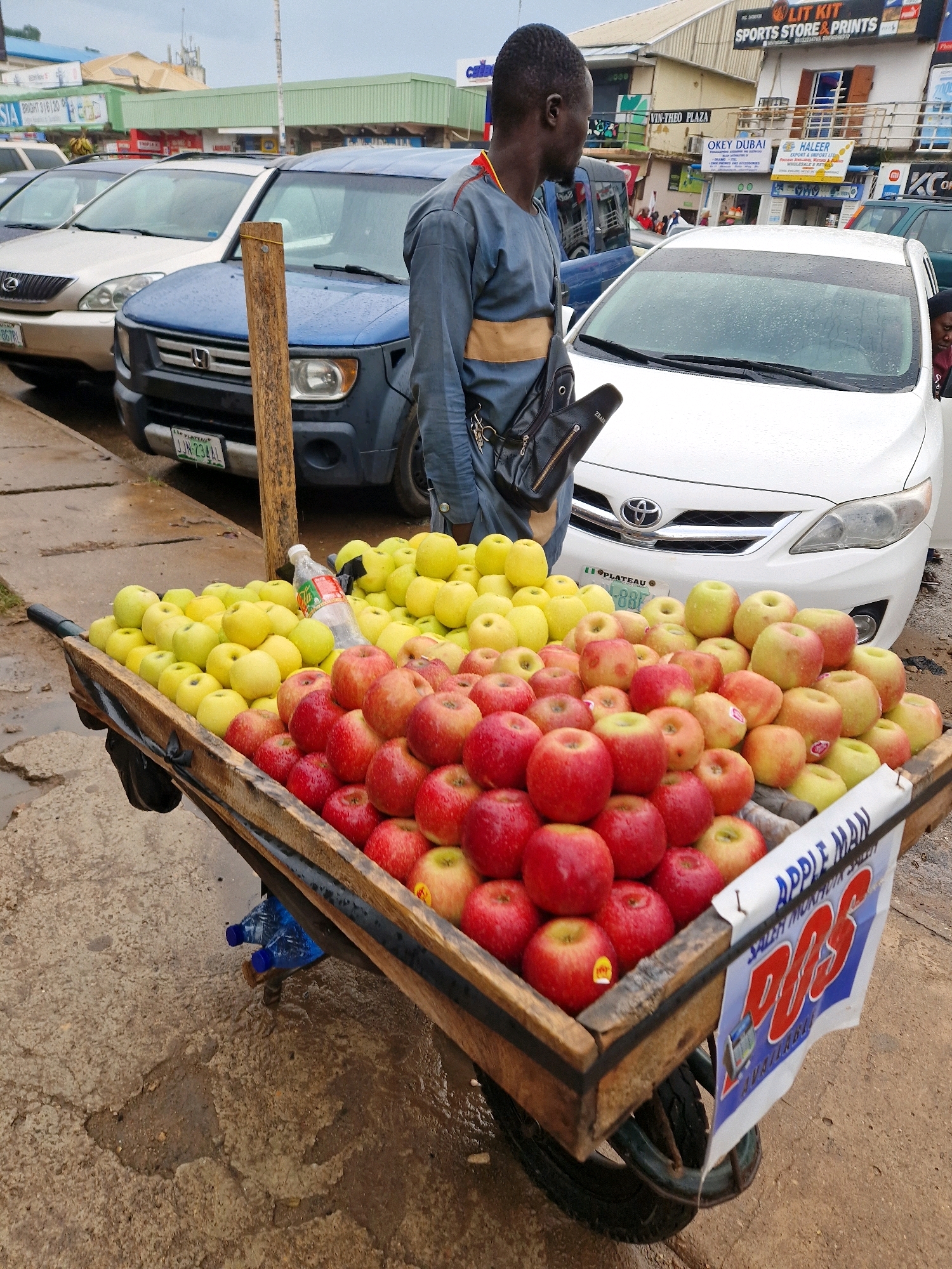 An eye-level, medium shot captures a street vendor's cart laden with apples and other fruit, parked on a wet, concrete surface in Jos, Nigeria. The vendor, a man wearing a grey traditional outfit with a yellow sash and a black crossbody bag, stands beside the cart. The cart, made of wood and metal, is overflowing with piles of red and yellow apples. A plastic bottle of water sits amongst the fruit. Behind the cart, several vehicles are parked, including a blue SUV and a white sedan with a license plate reading "PLATEAU 88F." In the background, a row of buildings with shops and signage, including "LIT KIT SPORTS STORE & PRINTS" and "OKEY DUBAI," line the street. The overall atmosphere suggests a typical market scene in a Nigerian city, possibly during or after rainfall, indicated by the wet ground and the reflections on the car windshields.