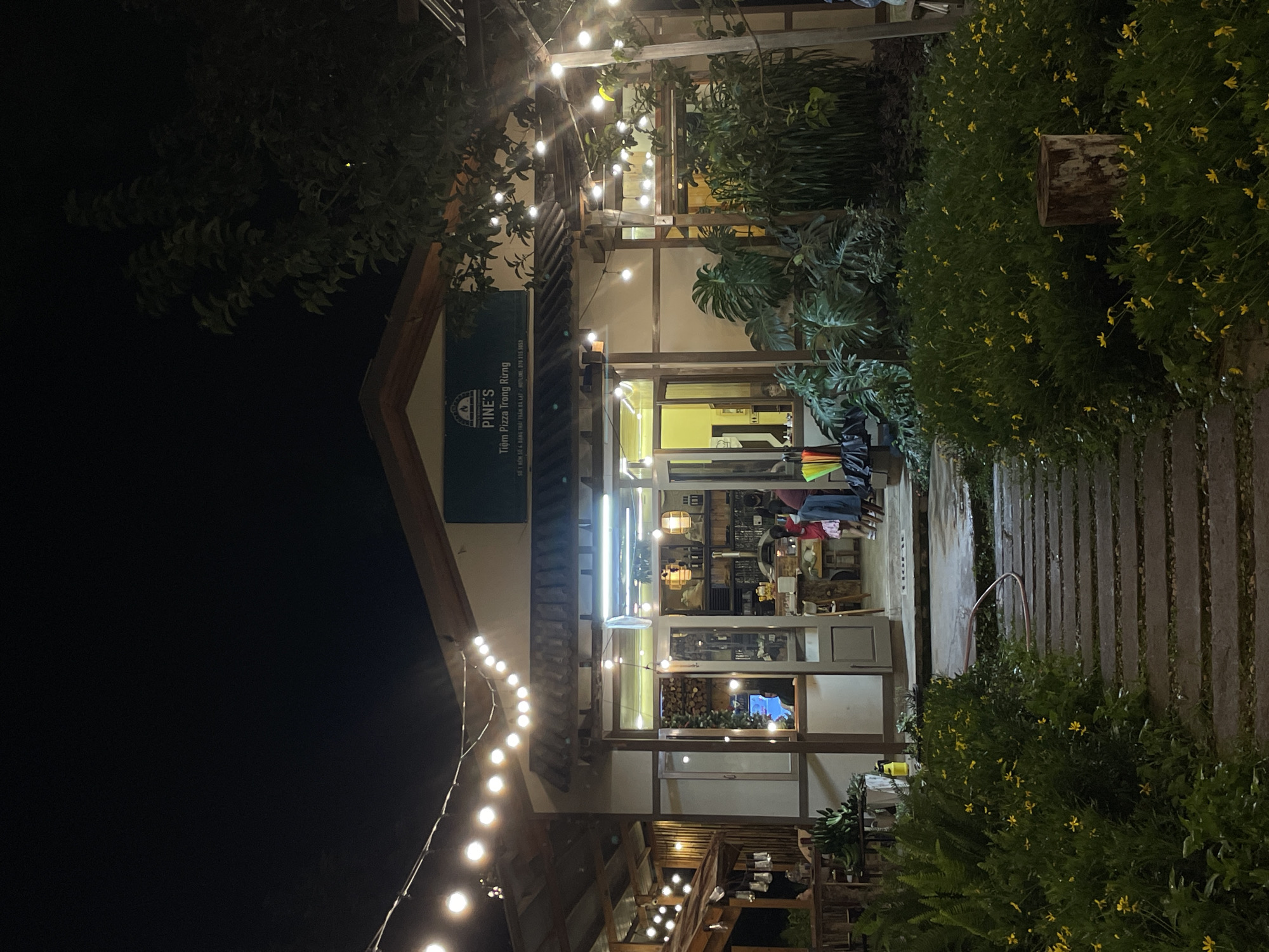 The image shows a nighttime view of Pine's Pizza, a restaurant located in Da Lat, Vietnam. The restaurant appears to be a single-story building with a rustic, wooden-accented facade. String lights are draped across the exterior, illuminating the entrance and outdoor seating area. The entrance features large glass doors, offering a glimpse into the interior where tables, chairs, and shelving units stocked with items are visible. Several patrons, including children, are present inside and near the entrance. A pathway made of stepping stones leads to the restaurant, flanked by lush greenery and small yellow flowers. The surrounding environment is dark, indicating nighttime, with trees visible on the right side of the frame. A sign above the entrance reads "PINE'S Tiệm Pizza Trong Rừng," which translates to "Pine's Pizza in the Forest."