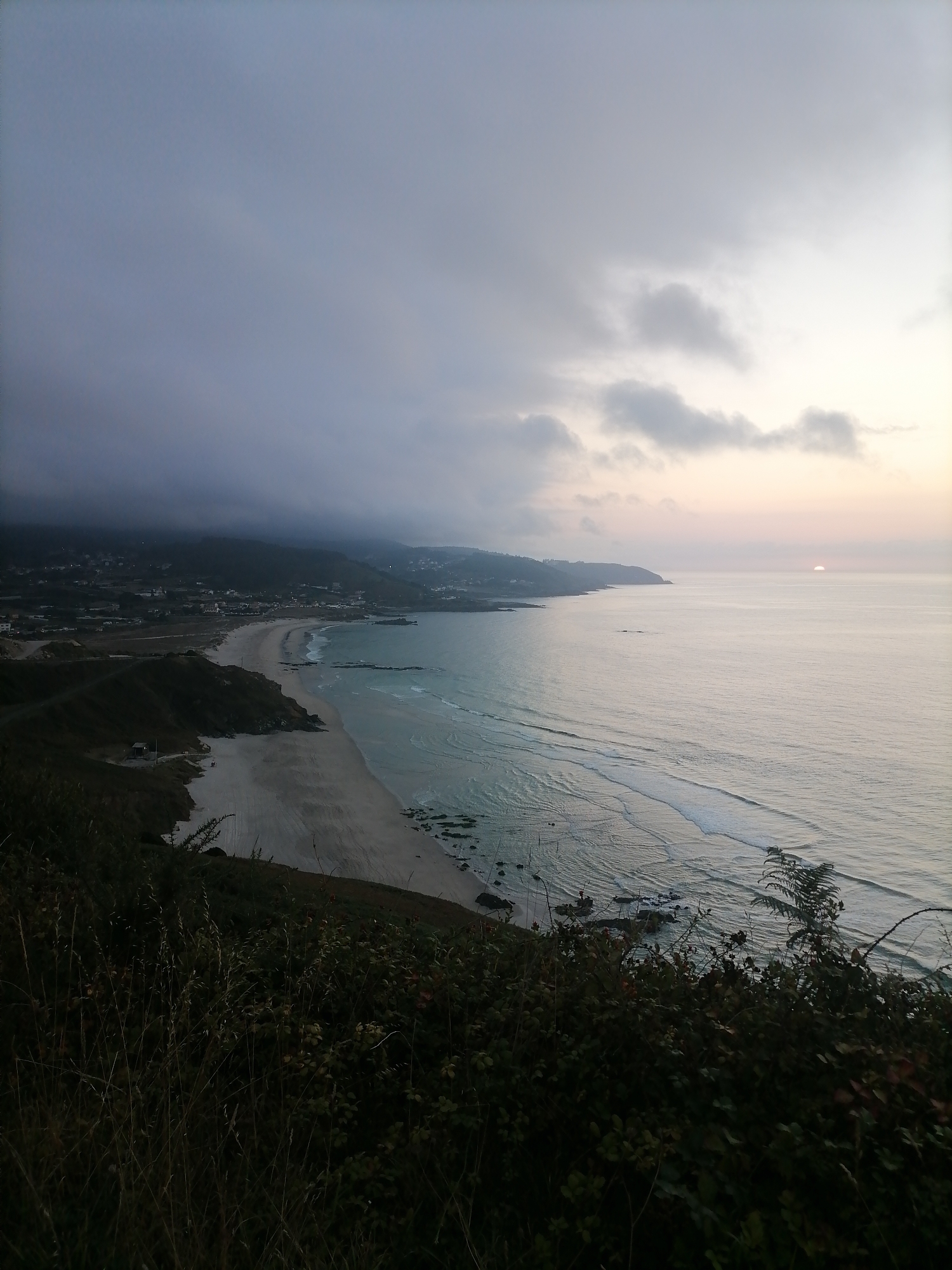This is an eye-level, wide-angle, outdoor photograph of the coastline in Arteixo, Spain. The image depicts a panoramic view of a beach with the ocean to the right, and a hillside with vegetation in the foreground. The sky is overcast with some lighter patches indicating the presence of the sun, which is partially visible on the horizon. The ocean is calm with gentle waves rolling onto the sandy beach. Buildings and houses are visible on the distant hillsides. The foreground is dominated by dark green foliage and dry grasses.