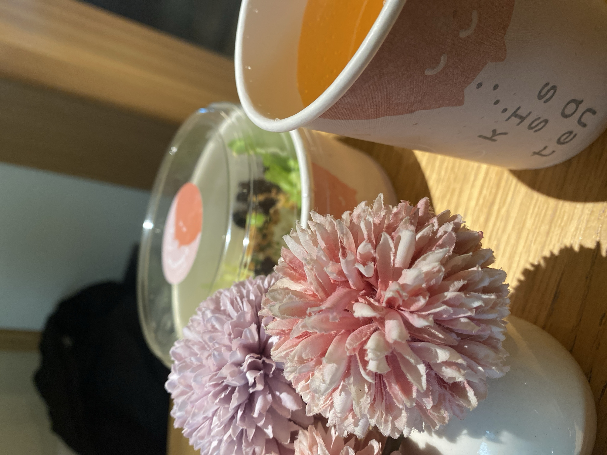 The image shows a table setting with a cup of orange juice, a salad in a clear container, and a vase with pink and lavender flowers. The objects are arranged on a wooden surface, possibly a table, with sunlight illuminating the scene. The text on the cup is only partially visible, reading "K..." and "...OC FRESH SO". No people are visible. There is no indication in the image that can link it to Krasnodar, Russia.