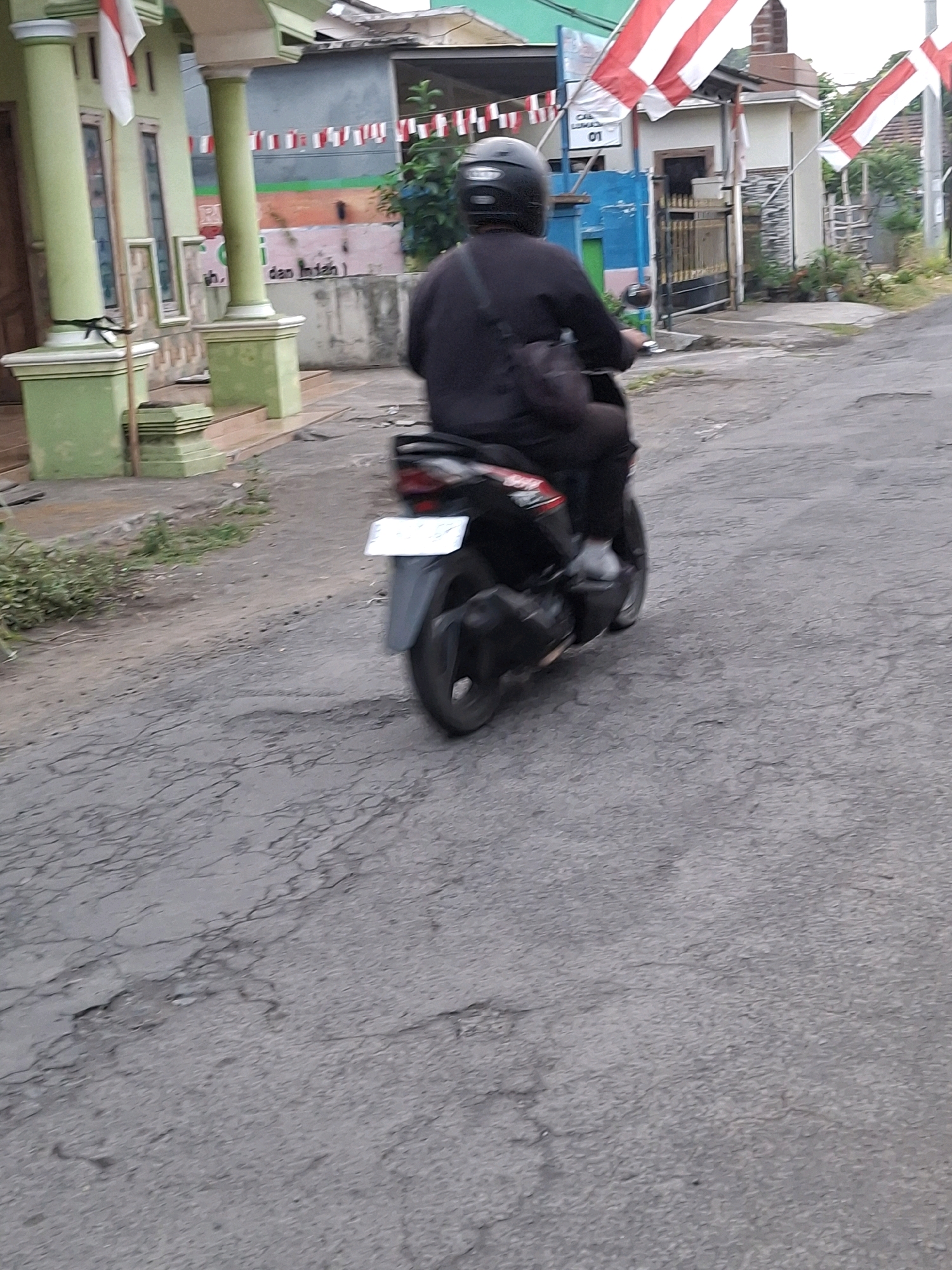 A person wearing a black helmet and dark clothing is riding a black motorcycle on a cracked asphalt road. The motorcycle has a white license plate. In the background, there is a light green building with a white pillar and a red and white flag. There are also strings of red and white flags and a blue and green wall with text that reads "CAL LEMAAN 01". The scene appears to be a street in Lumajang, Indonesia.