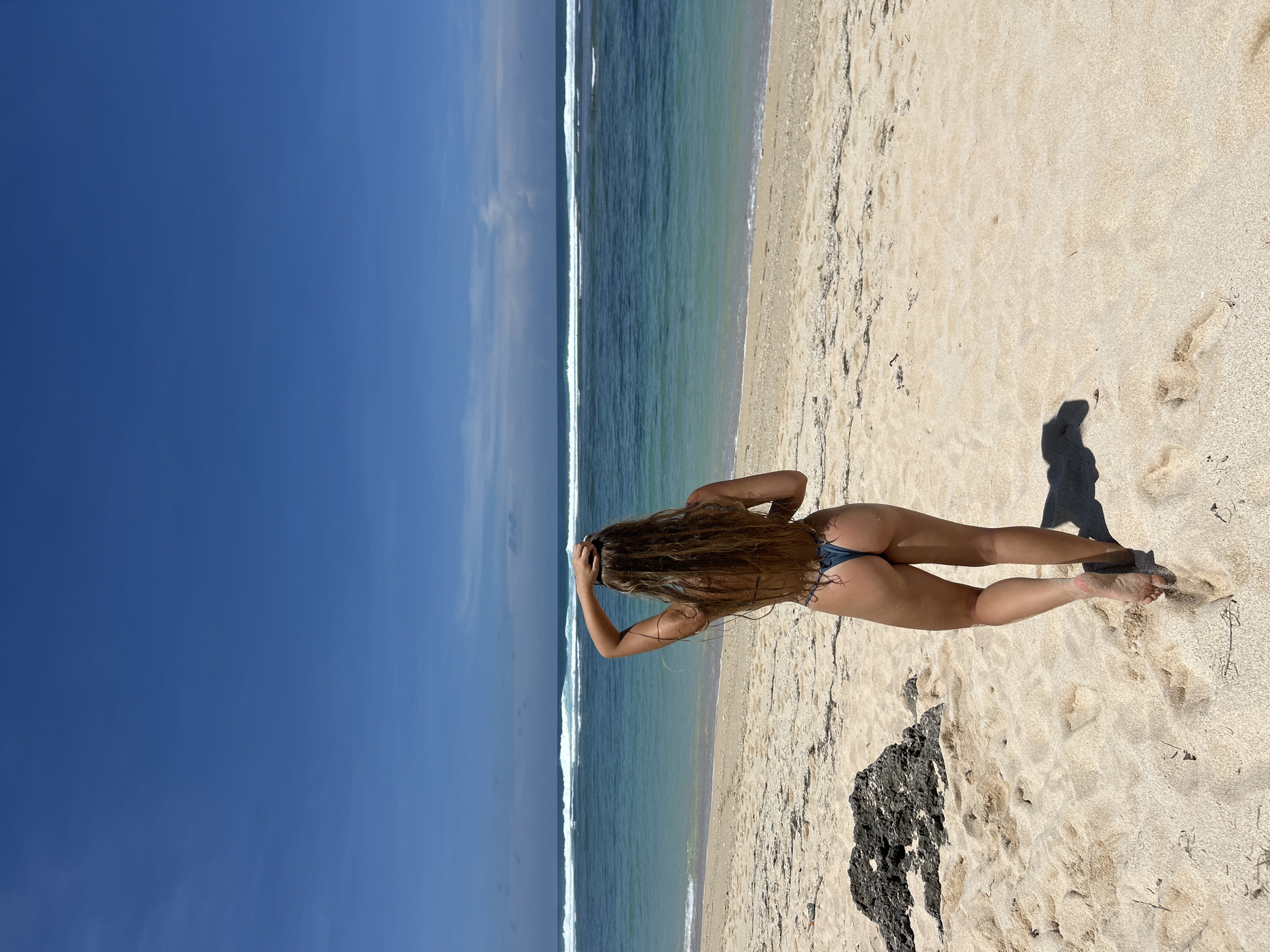 A woman stands on a beach in Kuta Selatan, Indonesia. She faces the ocean, her back to the viewer. She has long, wavy brown hair and wears a blue bikini bottom. Her hands are on her head. The beach has light-colored sand. A dark, rocky area is to the right. The ocean has light waves. The sky is bright blue.