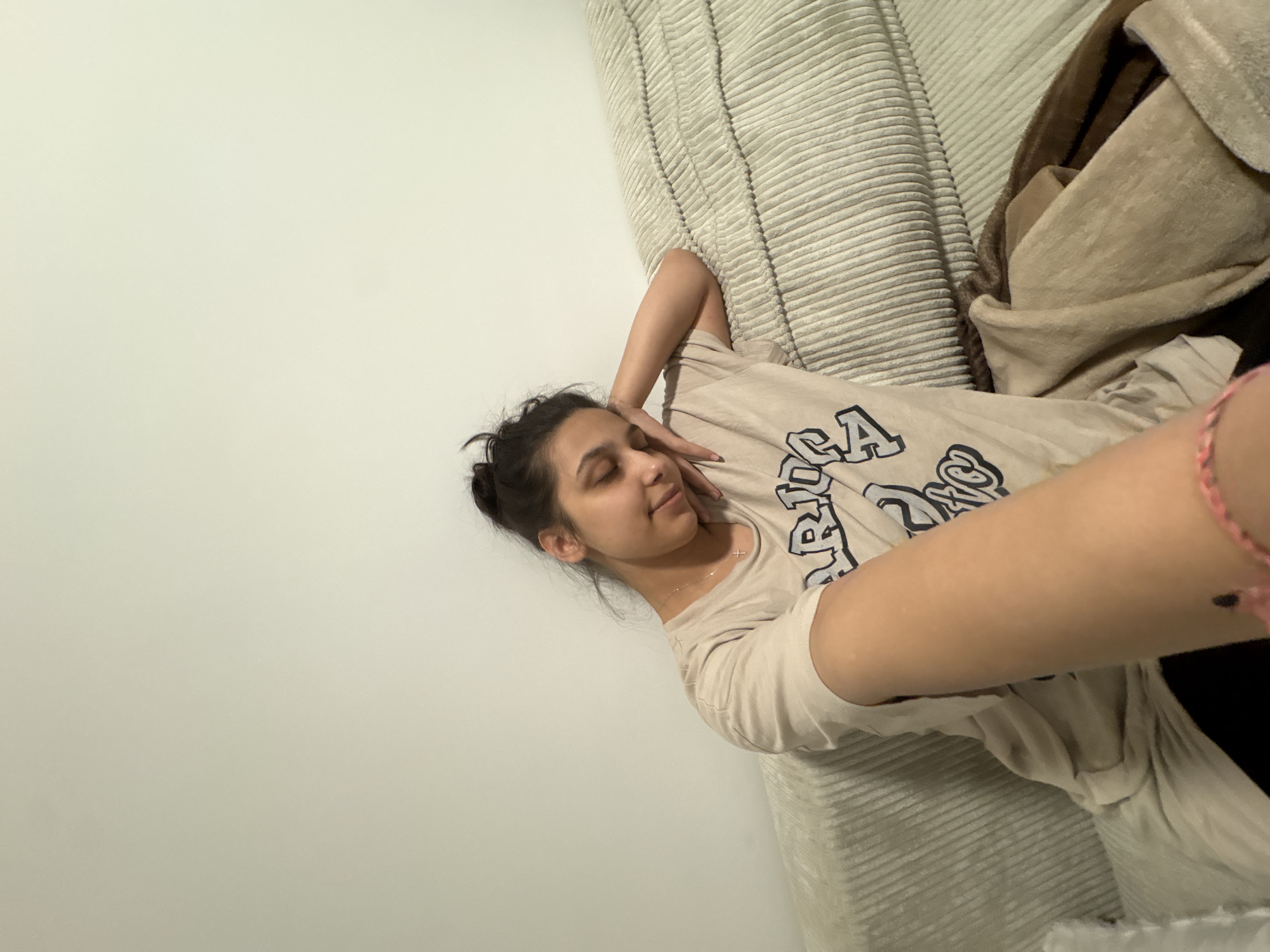 The image shows a young woman with dark hair pulled up in a bun, leaning her head on her hand while sitting on a couch. She is wearing a light-colored t-shirt with a graphic print. The couch appears to be made of a textured fabric. The background is a plain, light-colored wall. The overall impression is one of relaxation or rest. The image does not provide any information about the location in Mönchengladbach, Germany.