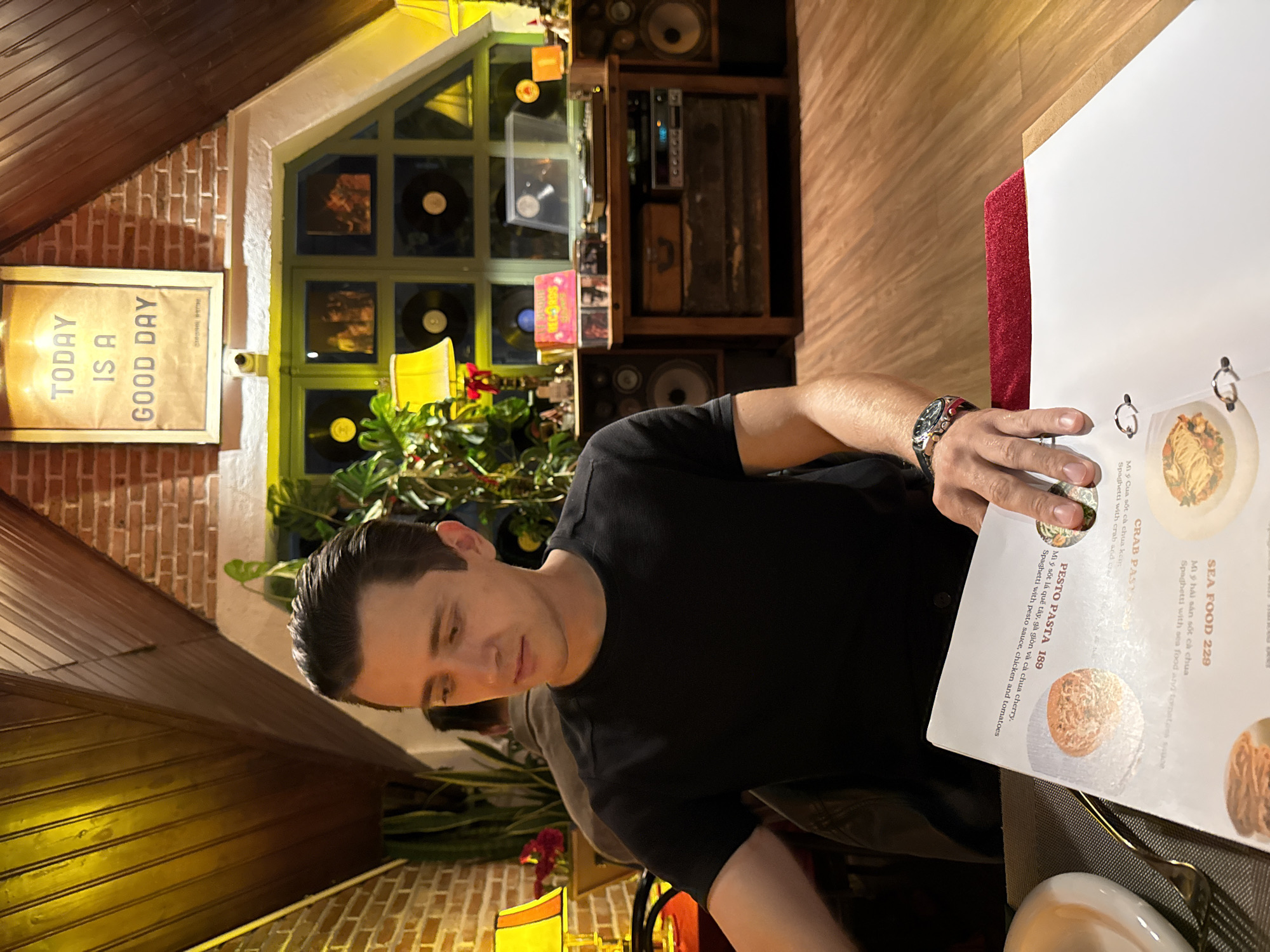 A man in a black t-shirt sits at a table in a restaurant, looking at a menu. The menu is in Vietnamese, and shows images of pasta dishes, including "Sea Food 229," "Pesto Pasta 189," and "Crab Pasta." The restaurant's decor includes brick walls, wooden furniture, records, and plants. A sign reads "Today is a Good Day." The restaurant is likely in Lâm Viên, Đà Lạt, Vietnam.
