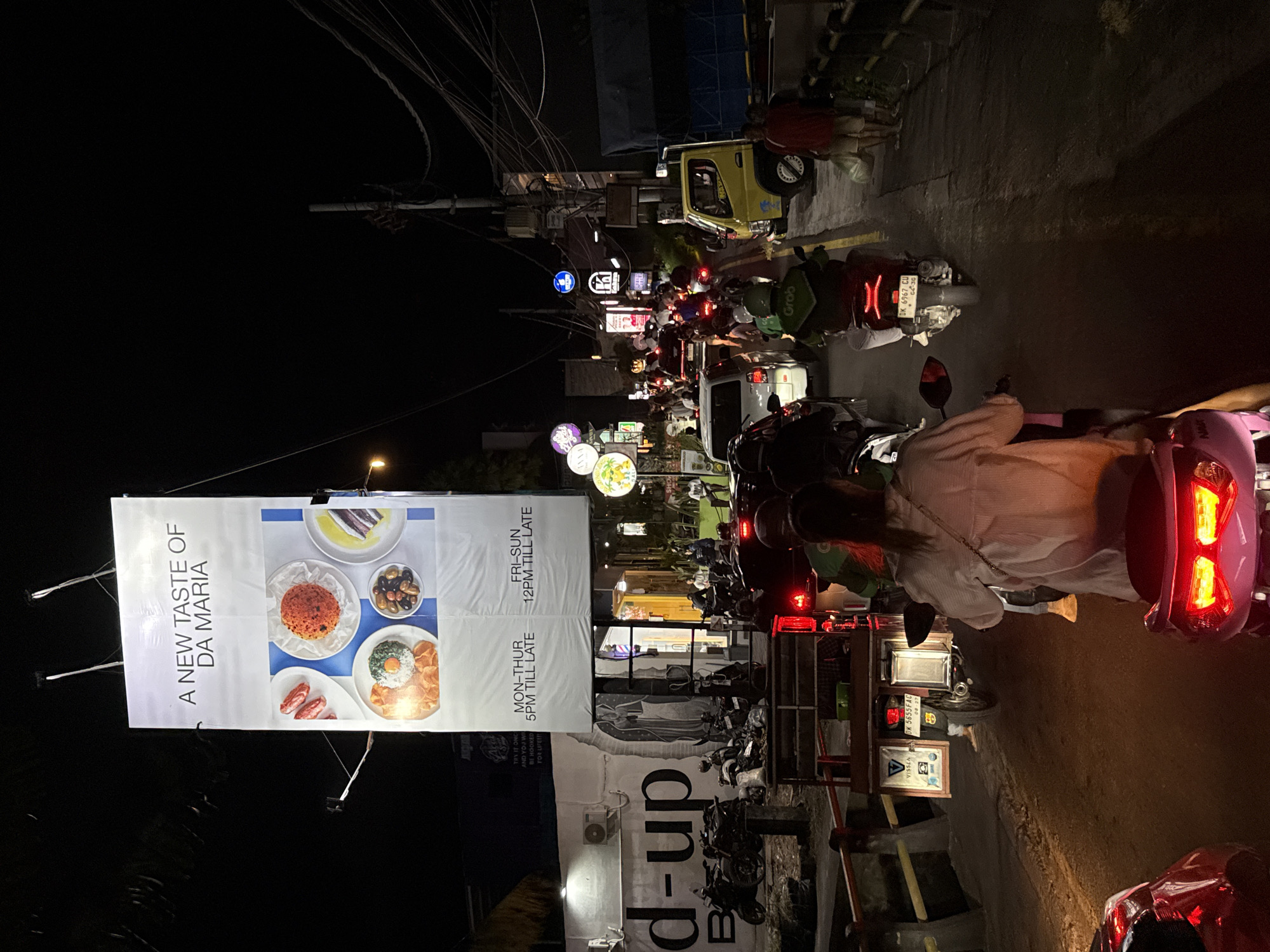 The image depicts a night scene in North Kuta, Indonesia, characterized by a busy street with numerous motorbikes and vehicles. A prominent banner advertises "A New Taste of Da Maria" with business hours "MON-THUR 5PM TILL LATE, FRI-SUN 12PM TILL LATE." The street is illuminated by various signs and vehicle lights, creating a vibrant atmosphere. People are riding motorbikes, contributing to the lively street scene. A "Grab" motorbike is visible. In the foreground, a scooter rider is shown, and other motorbikes are in the line.