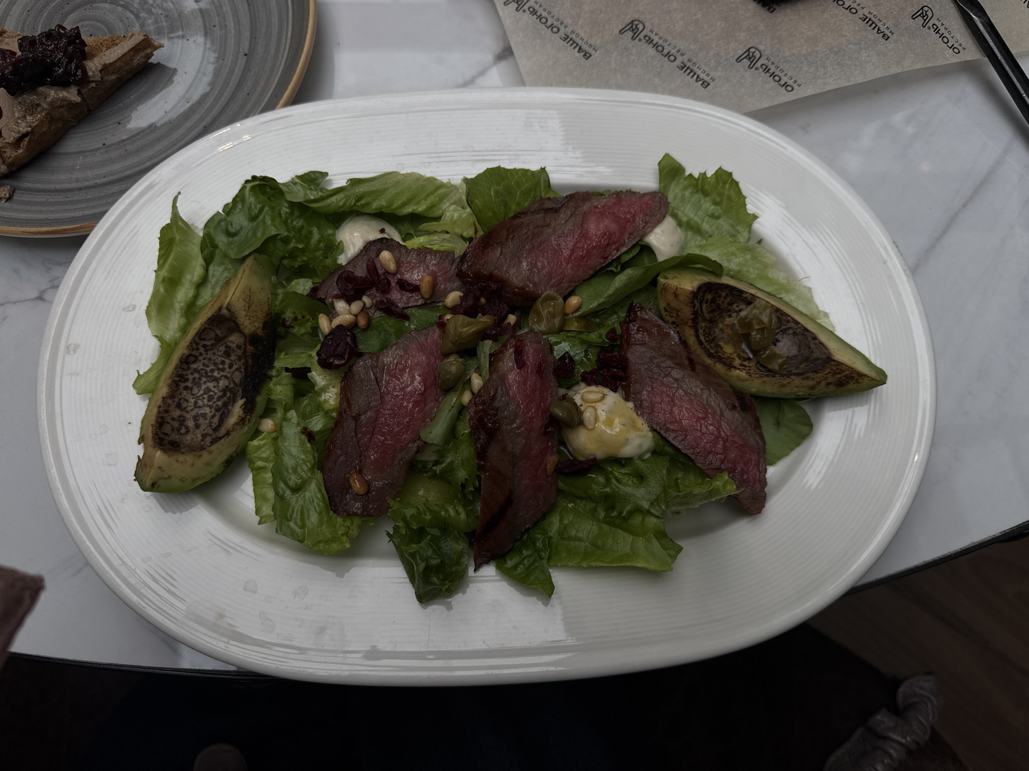 The image displays a top-down view of a meal served in an indoor restaurant setting in Izhevsk, Russia. A large white rectangular plate, centrally positioned, holds a salad. The salad base consists of green leaf lettuce, topped with approximately six slices of medium-rare cooked steak. Two halves of grilled or roasted avocado, showing darkened, slightly charred interior surfaces, are placed on opposite ends of the plate. Interspersed within the salad are white dollops of creamy dressing, scattered light-colored pine nuts, small dark red pieces (possibly dried cranberries), and small green capers or pickled peppers.
To the right, partially visible behind the main plate, a smaller round grey plate holds a triangular slice of toast. This toast is spread with pâté and garnished with a dark, berry-like preserve. On the right side of the table, a beige paper napkin or placemat is visible, bearing repeated black text and a logo. The text, in Cyrillic, reads "ВАШЕ ОГОНЬ" (Your Fire/Flame) and "МЯСНОЙ РЕСТОРАН" (Meat Restaurant), indicating the establishment specializes in meat dishes. A stylized "O" or flame logo accompanies the text.
A person's right hand, with a dark sleeve, is partially visible in the upper left corner of the frame, positioned as if reaching for or about to interact with the food. The table surface is a light-colored material, possibly marble or stone, with subtle veining. A dark, indistinct object, possibly footwear or clothing, is partially visible in the bottom left.