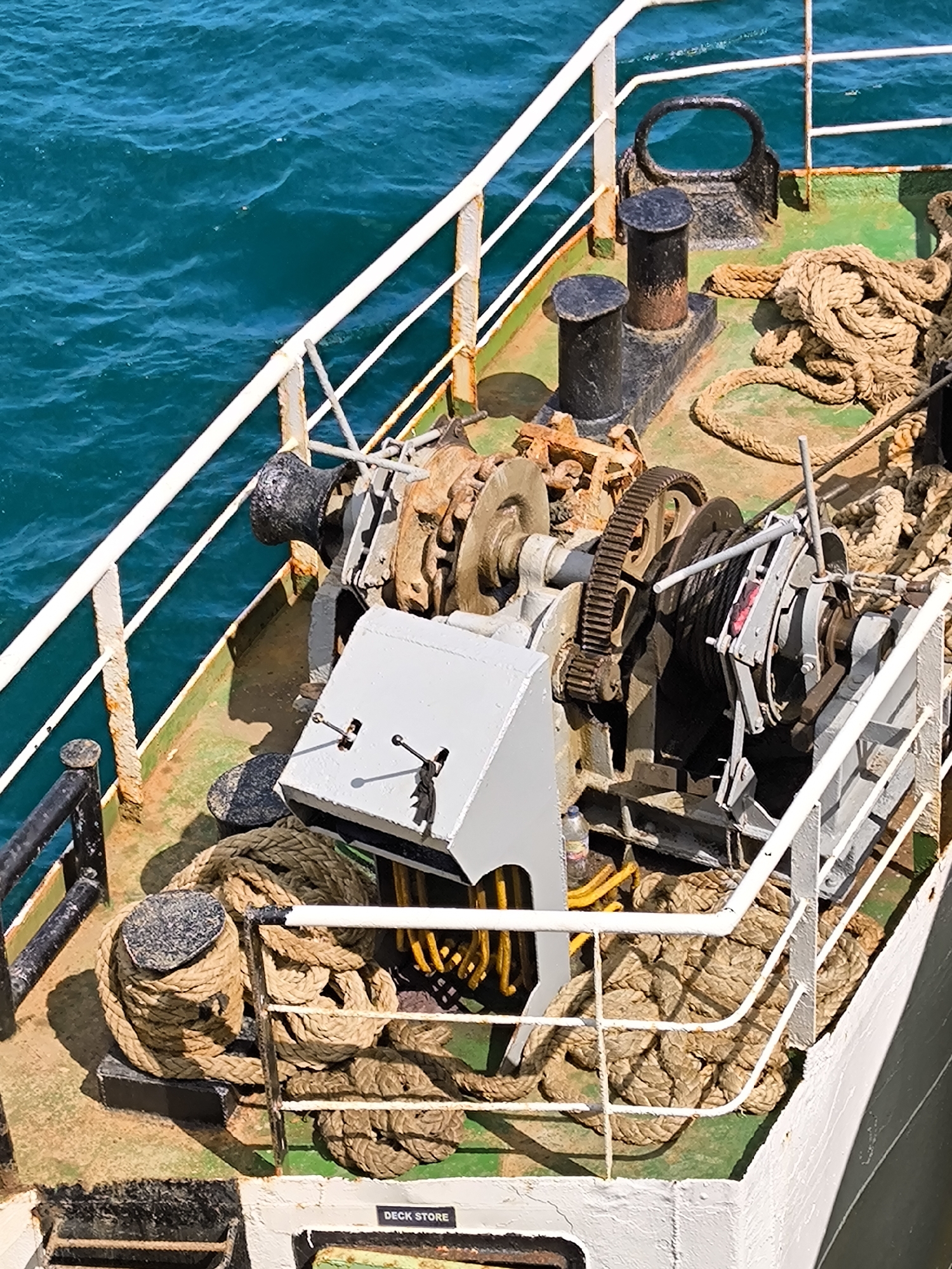 A high-angle, outdoor shot captures the working deck of a ship or ferry alongside vibrant turquoise-blue water, likely in Lembar Selatan, Indonesia. The scene is brightly lit by daylight, casting distinct shadows and indicating clear, sunny weather.
The deck, painted in a worn green and brown, features various maritime equipment. In the central area, a robust winching system is prominent, characterized by large gears, spools, and manual levers, suggesting its use for anchoring or mooring operations. Nearby, black cylindrical bollards and a larger U-shaped bitt are affixed to the deck, serving as points for securing ropes.
Numerous thick, coiled ropes are scattered across the deck; some are neatly coiled around a bollard, while others lie in loose heaps. A white metal railing, showing signs of rust and wear, runs along the deck's edge, separating it from the open water. No people are visible, emphasizing the functional nature of the ship's hardware.
Other details include a small plastic bottle tucked near some yellow cables or hoses beside the winch. The machinery and deck surfaces display visible signs of age, wear, and rust, indicating a well-used working vessel. The text "DECK STORE" is clearly visible on a white panel towards the bottom right of the image, labeling a storage area. The clear, rippling water underscores the vessel's maritime environment.