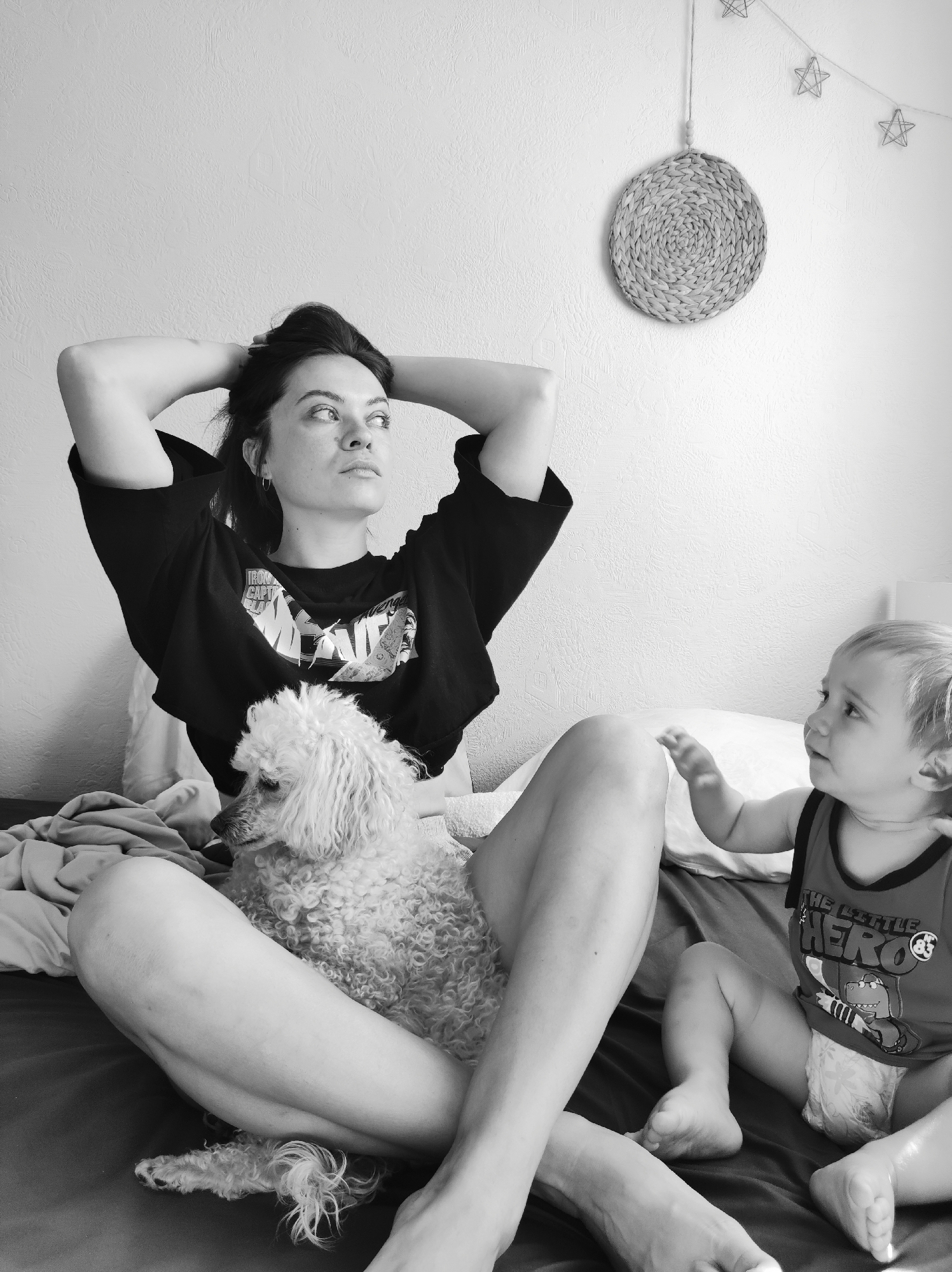 The image, in black and white, shows a woman sitting on a bed with a poodle in her lap. A baby sits on the bed to her right. The woman has dark hair pulled back, a dark t-shirt, and her hands behind her head. The poodle is light-colored with curly fur. The baby wears a tank top with a graphic and a diaper. The bed has a dark-colored sheet and pillows behind the woman. On the wall behind the bed is a woven, circular decoration and a string of star-shaped lights. The setting is likely indoors, possibly a bedroom. There is no information confirming the location as Chaikovsky, Russia.