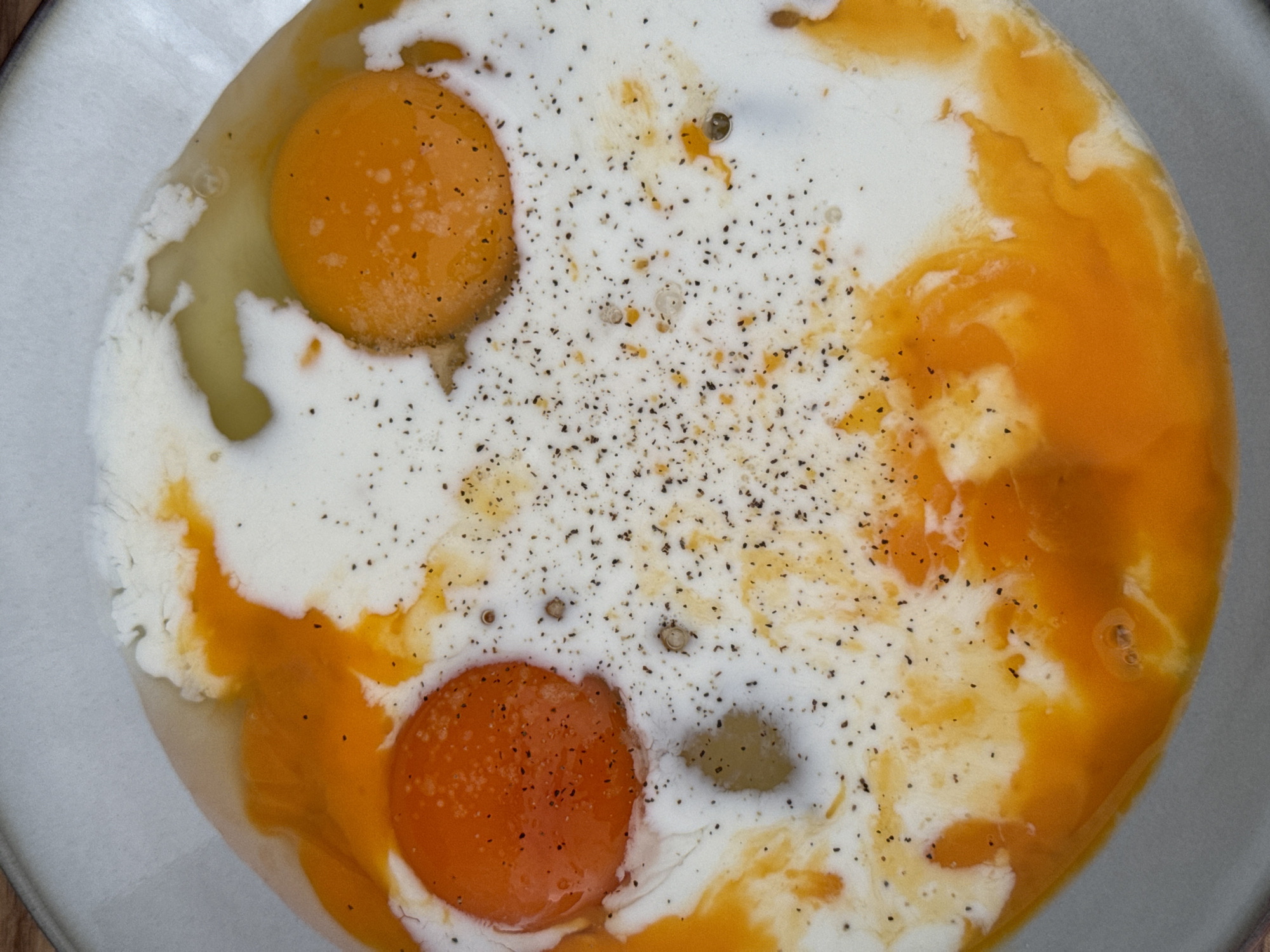 A top-down view of a light gray bowl containing two raw egg yolks, some egg whites, and a white liquid, possibly milk or cream. Black pepper is sprinkled over the mixture. The bowl appears to be sitting on a wooden surface. No information is available relating to Bjeliši, Montenegro.