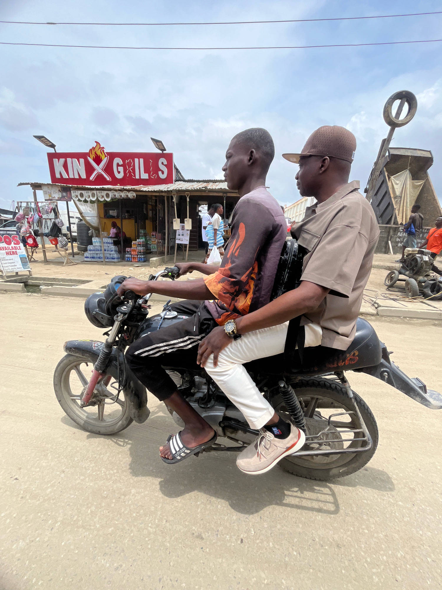 Two young men are riding on a motorcycle down a dirt road in what appears to be an outdoor market in Ado, Nigeria. The man in front, wearing a brown cap and shirt, is driving. The man behind him, wearing a patterned shirt and white pants, is a passenger. They are both looking forward. In the background, a stall with a sign that reads "KING GRILLS FOODER" is visible, along with other market stalls and people. The sky is overcast.