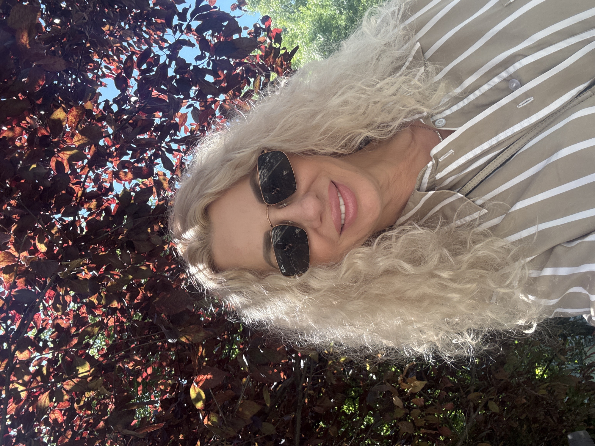 A medium shot shows a blonde woman with curly hair, wearing sunglasses and a striped button-down shirt. She is outdoors, with red leaves and blue sky visible behind her, suggesting a park or garden setting. The background foliage indicates it is likely daytime and possibly autumn. The image captures a close-up of the woman's face and upper torso, with her sunglasses reflecting some of the surroundings.