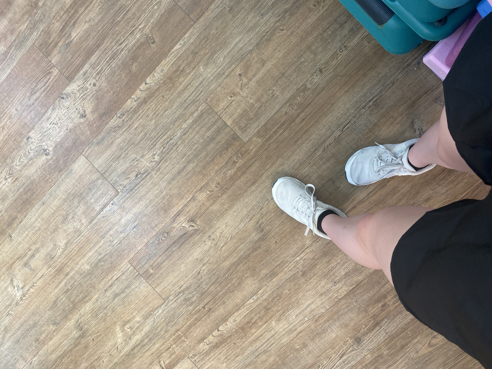 An overhead perspective shot shows a person's legs and feet, clad in white athletic shoes and black shorts, standing on a wooden floor. The floorboards are laid diagonally, with visible wood grain and knots. To the right of the frame, a teal aerobic step and a pink block are partially visible. The scene suggests an indoor setting, possibly a gym or fitness studio.