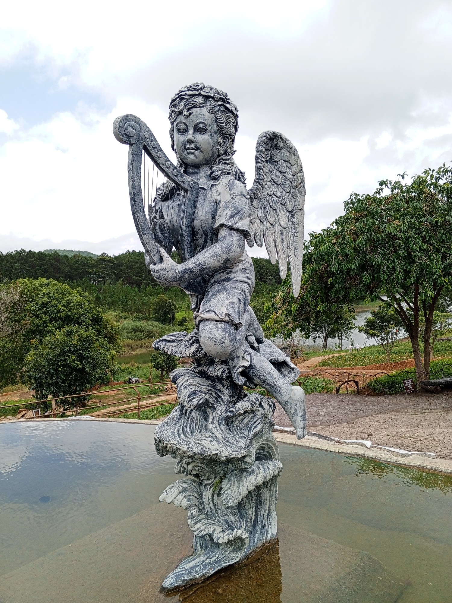A statue of a winged cherub holding a harp is positioned in the foreground, partially submerged in a shallow pool of water. The cherub, made of a gray, textured material, is seated on a base that resembles waves or clouds. It has curly hair, prominent facial features, and detailed wings. The background reveals a natural landscape with lush green trees, a body of water, and a cloudy sky. In the middle ground, a pathway made of interlocking stones is visible, along with a wooden bench and a fence. The overall scene suggests an outdoor garden or park setting. The statue is situated at the edge of a pool or water feature, likely part of a decorative landscaping element.
