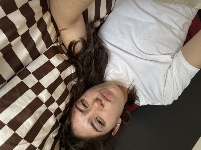 A person is lying on their back on a bed. They are wearing a white t-shirt. Their head is angled towards the viewer, with dark hair spread around them. The bed has a checkerboard patterned comforter and a burgundy pillow. The background is black. The person has fair skin, dark eyes, and a small nose ring.
