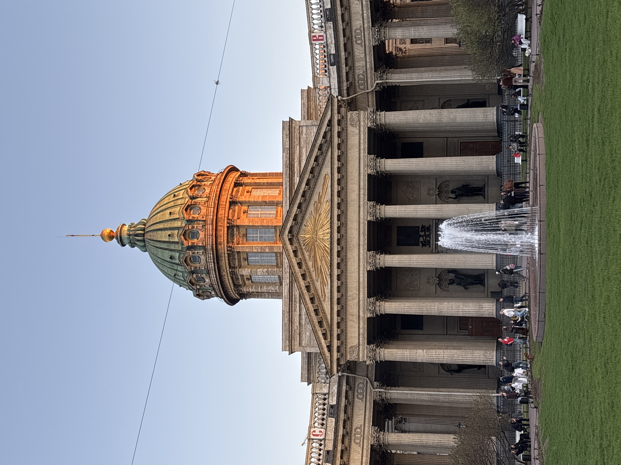 The image depicts the Kazan Cathedral in Saint Petersburg, Russia. The architecture is neoclassical, featuring a large dome and a colonnade. A fountain is in the foreground, and people are sitting and walking nearby. The environment suggests a public space, possibly a park or square.