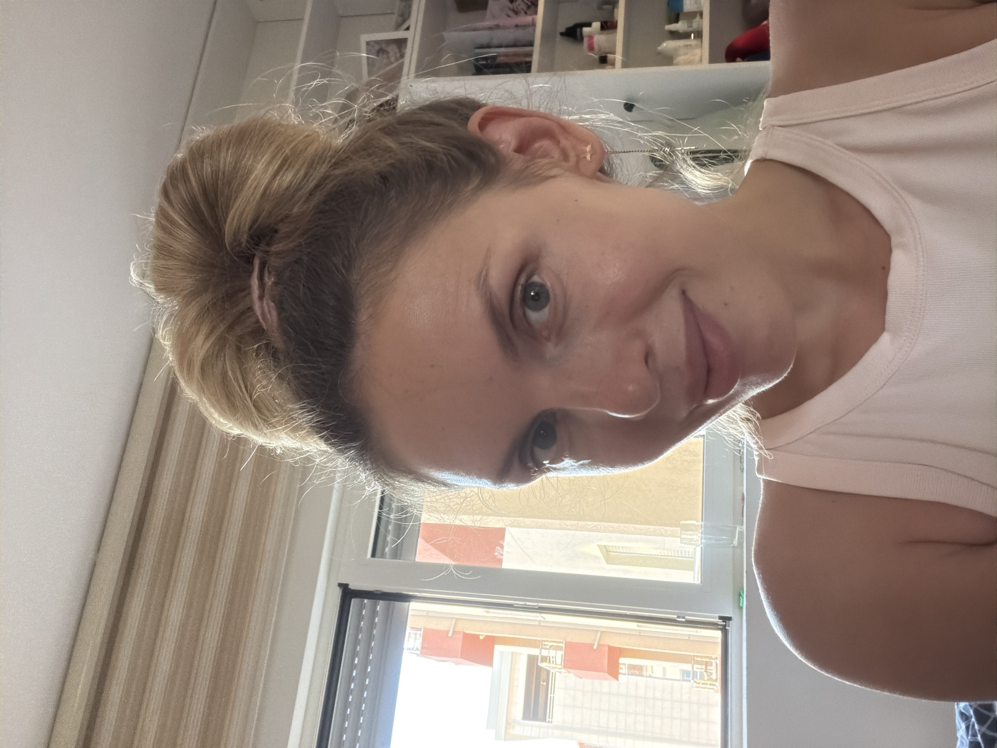 A close-up, over-the-shoulder shot shows a woman with blonde hair pulled up in a bun, facing the camera. She is wearing a light pink tank top. Her left shoulder is visible in the foreground. Behind her, a window reveals a city view with buildings and balconies, consistent with the architecture found in Budva, Montenegro. The lighting suggests it is daytime, with sunlight streaming in from the window. To the right of the woman, shelves are visible, stocked with various items.