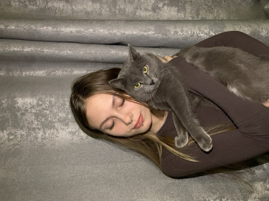 The image depicts an indoor setting, reportedly in the city of Foki, Russia. A female subject, appearing to be in her late teens or early twenties, with long, light brown hair, is holding a domestic cat. She wears a dark brown long-sleeved garment. Her visible facial piercings include a silver stud in her right nostril and two small silver studs positioned horizontally below her lower lip. Her eyes are closed, and she has a subtle smile. She cradles a medium-sized cat with dense, grey fur and prominent yellow-green eyes. The cat's front paws are extended across her left arm, and its gaze is directed forward towards the viewer. The background consists of a textured, light grey material, possibly a curtain or decorative wall covering, exhibiting vertical folds and mottled variations in tone.