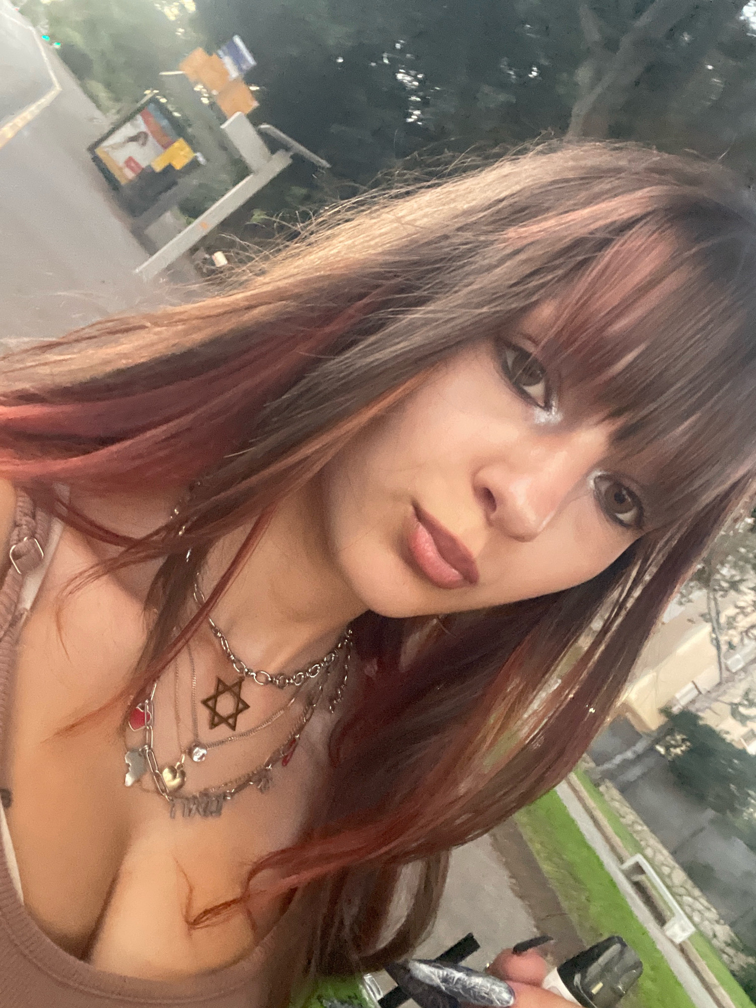 The image is a close-up selfie of a young woman with reddish-brown hair and bangs. She is wearing a brown top and several necklaces, including one with a Star of David pendant and another with Hebrew lettering. Her makeup includes prominent eyeliner. The background is out of focus, showing a street with trees and a sign that appears to be in Israel, possibly Raanana. The lighting suggests it is late afternoon or early evening. A vaping device is visible in the lower right corner, held by the woman's hand which has long, dark nails.