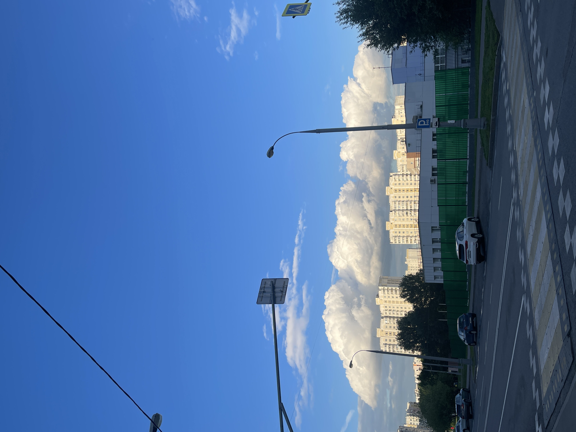 This image depicts a street scene in Moscow, Russia. The primary subjects are buildings, a street, a crosswalk, a lamppost, and several cars. The sky is a vibrant blue with scattered white clouds, suggesting it is daytime, likely late afternoon given the lighting. The presence of a crosswalk and streetlights indicates an urban environment.
In the foreground, a portion of a paved road with a yellow and white striped crosswalk is visible. A lamppost with a curved arm stands on the side of the road. Further back, several cars are parked or driving along the street. One white SUV is clearly visible, and a dark-colored car is behind it.
Behind the street, there are a series of multi-story buildings. The architecture suggests a residential or commercial area typical of Russian cities. Some buildings have a light-colored facade, while others are clad in green panels. Trees are interspersed between the buildings and along the roadside.
A road sign is visible on a pole above the lamppost, though its details are not discernible. Another sign, a blue parking sign with a white "P" and a clock symbol, is mounted on a pole near the green fence. A pedestrian crossing sign, a yellow diamond with a black silhouette of people, is also visible in the upper part of the frame.
The scene is bathed in sunlight, casting soft shadows, indicating a pleasant day. There are no people visible in the frame, making it a quiet, almost deserted street scene. The overall impression is one of an ordinary urban day in Moscow.