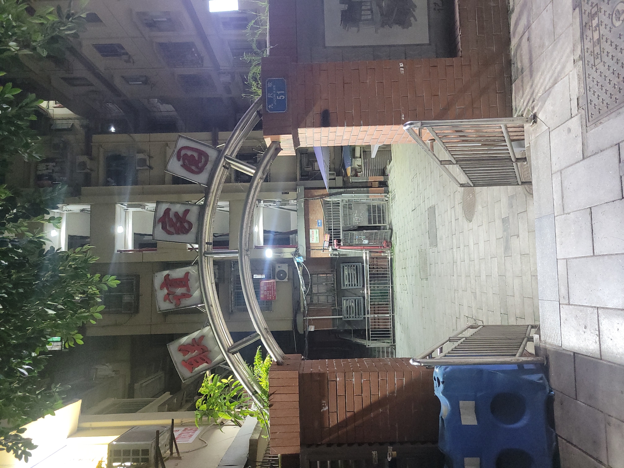 The image displays an urban residential scene at night, viewed from an alleyway or entrance. The overall atmosphere is quiet, illuminated by artificial lights.
In the foreground and center, a prominent, curved metallic archway spans the path. Three square signs hang from this archway, each featuring a stylized red Chinese character on a white background. From top to bottom, these characters appear to be "园" (yuán), "悦" (yuè), and "居" (jū), likely forming "园悦居", which translates to "Garden Joy Residence" or similar, suggesting the name of a residential complex. Green foliage from trees or shrubs is visible on the left and near the archway's base.
To the right, a reddish-brown brick wall supports the archway. A blue rectangular street sign is affixed to this wall, clearly displaying the Chinese characters "九尺坎" (Jiǔchǐ kǎn) and the number "51". Metal railings extend along the right side, bordering a walkway or steps. Another set of metal railings is visible further back, near an entrance or gate. In the bottom right corner, a large blue object, possibly a barrier or bin, is present.
The ground is paved with light-colored, rectangular tiles, and several manhole covers are visible. In the background, multiple-story residential buildings rise, their windows glowing with internal lights and exterior air conditioning units visible on their facades. No people or specific activities are depicted in the scene, which conveys a calm, nocturnal ambiance. The artificial lighting creates reflections on the paved ground and highlights the signage.