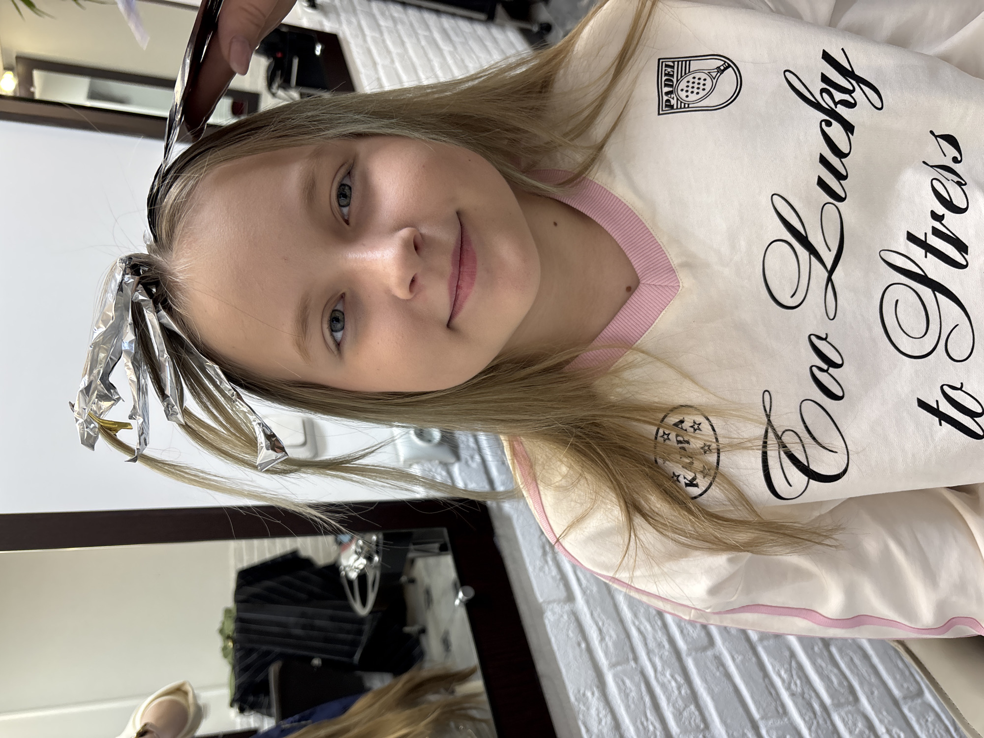 The image shows a fair-skinned young girl with light brown hair and blue eyes. She is smiling and looking at the camera. Sections of her hair are wrapped in aluminum foil, indicating a hair coloring process is underway. A hand holding a comb or brush is visible near her head, likely applying hair dye. She is wearing a light-colored sweatshirt with a pink neckline and black text that reads "Too Lucky to Stress". There's a "PADEL" logo on her upper chest.
In the background, a white brick wall can be seen, as well as a dark-framed mirror reflecting salon equipment, including a dark-colored storage unit. The setting is a hair salon, possibly in Izhevsk, Russia. The scene suggests someone is in the process of getting their hair dyed.