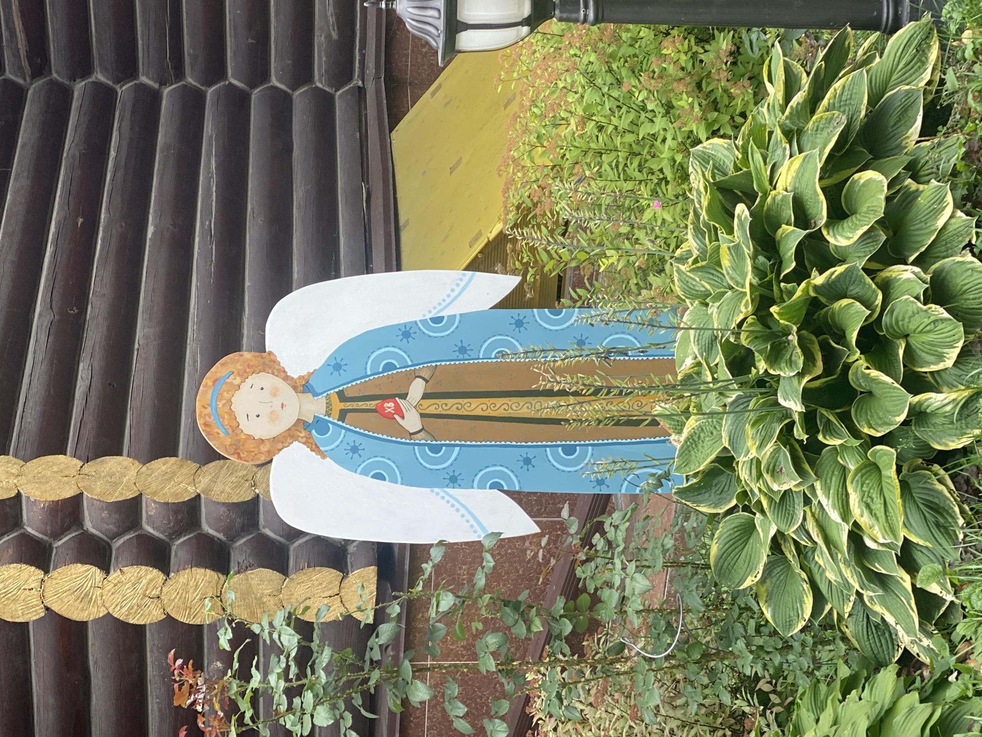 A wooden, cutout depiction of an angel stands in a garden in front of a log building. The angel has blonde curly hair, rosy cheeks, and is wearing a blue robe with circular patterns and a brown tunic. It holds a red heart with the Cyrillic letters "ХВ" in its hands. White wings extend from its back. The angel is surrounded by lush green hosta plants in the foreground and other greenery. In the background, the dark brown logs of a building are visible, with a section of a yellow roof to the right. A black lamp post with a white light fixture is partially visible on the far right. The overall impression is a decorative garden display.