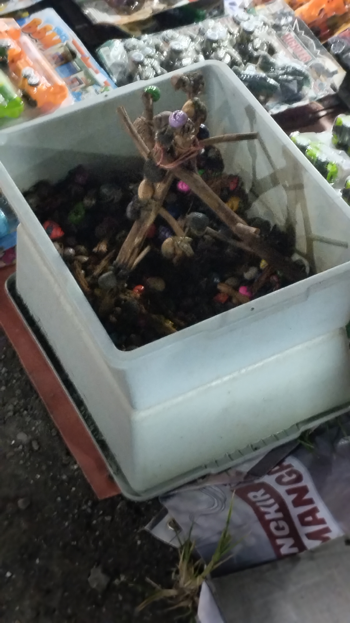 A white plastic tub filled with dark soil, small pebbles, and decorative elements, including sticks arranged in a tripod formation with beads of various colors attached, is positioned on a surface that appears to be a market stall. The tub is partially set against a backdrop of other market goods, some of which are packaged in clear plastic. The surface beneath the tub is dark and uneven, possibly concrete or packed dirt, with some scattered small stones. In the lower right corner, a portion of a banner or sign with text, possibly in Indonesian, is visible. The overall lighting suggests an outdoor setting, likely during the day.