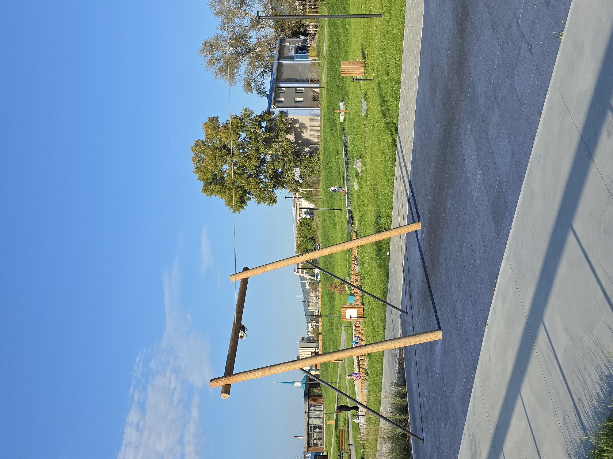 The image depicts a sunny day in a park in Miass, Russia. In the foreground, a large wooden swing set with two angled legs and a horizontal beam is prominent. The ground beneath the swing set is covered with dark, square rubber tiles. A gray concrete walkway borders the rubber tiled area.
In the midground, a grassy area stretches out with some children playing in a sandbox with wooden borders. A lone person is walking on the grass. Further back, a building with blue and gray siding and numerous windows is visible. Trees, including a large, leafy deciduous tree, are scattered throughout the park. The sky is a clear, bright blue with a few wispy white clouds. The scene suggests a public recreational area in the city.