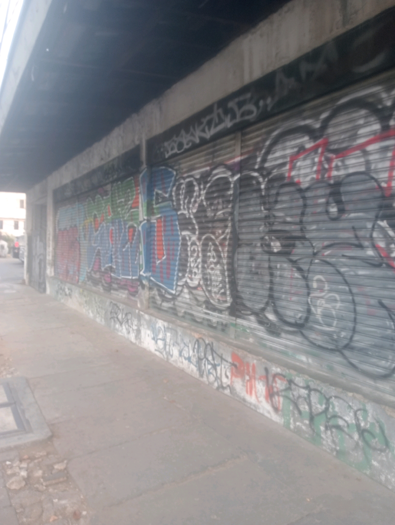 The image depicts a street-level view of a building's facade covered in graffiti. The facade appears to be composed of roll-up metal doors, also heavily graffitied with various tags and designs in multiple colors, including blue, red, green, and black. Above the roll-up doors, a concrete overhang or structure is visible, also showing some graffiti. The sidewalk in front of the building is paved with concrete and shows some signs of wear and tear, with debris scattered in places. To the left, a glimpse of buildings and street traffic is visible, suggesting an urban environment. The overall impression is one of urban decay and artistic expression through graffiti. The image does not provide enough visual information to definitively identify the location as East Jakarta, Indonesia.